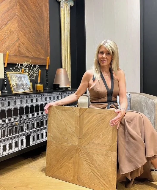 Mujer de cabello rubio con vestido largo marrón y negro, sentada con una mesa de madera en un ambiente decorativo con cuadros, lámparas y objetos decorativos.