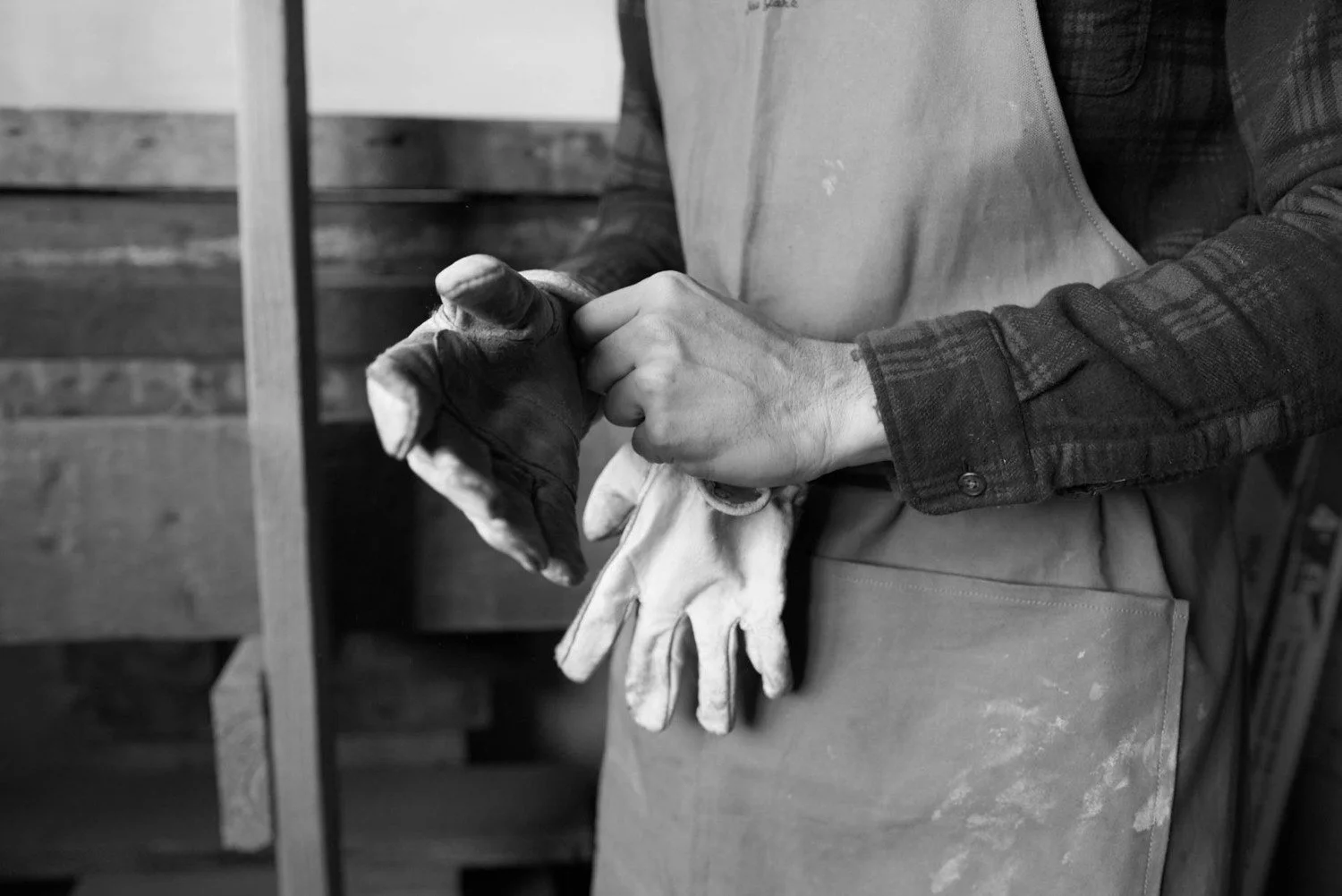Persona que usa un delantal y guantes manipula utensilios de trabajo en un taller o cocina, en un entorno de madera, en blanco y negro.