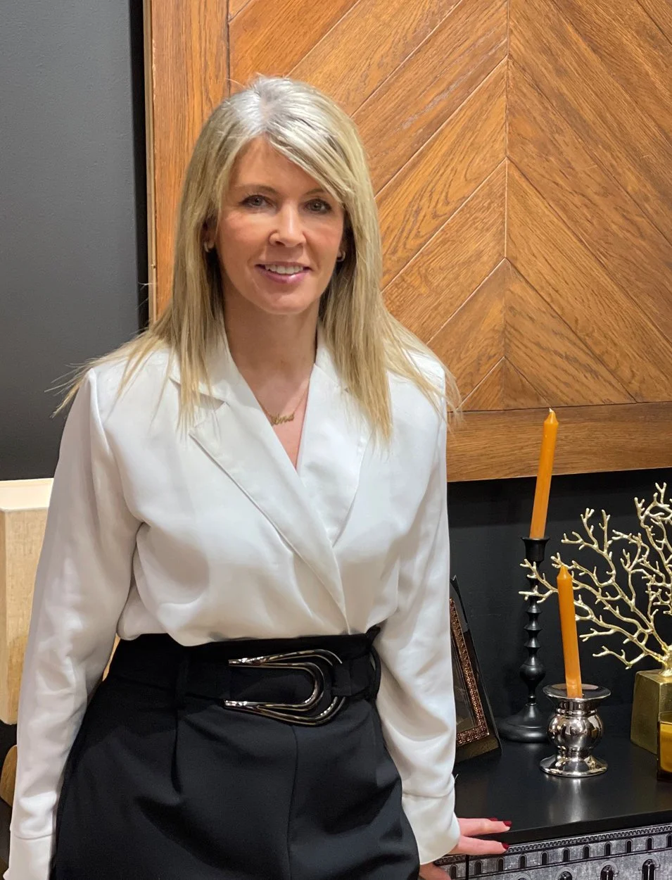Mujer de cabello rubio y largo, vestida con blusa blanca y falda negra, posando en un ambiente con decoraciones de candelabros y ramas doradas.