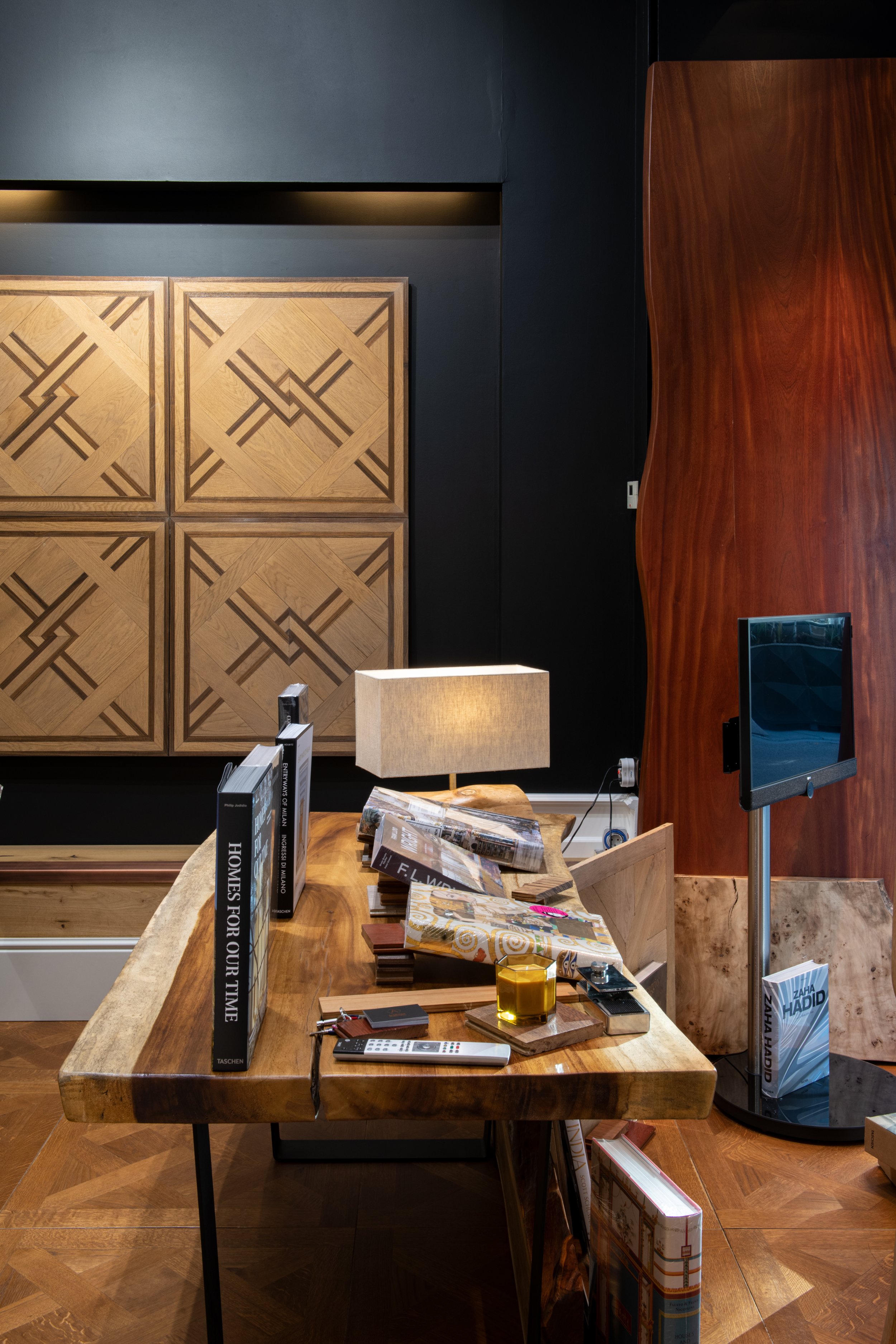 Mesa de madera con varios libros, revistas, y objetos personales en un ambiente moderno y elegante con decoraciones en madera y paredes oscuras.