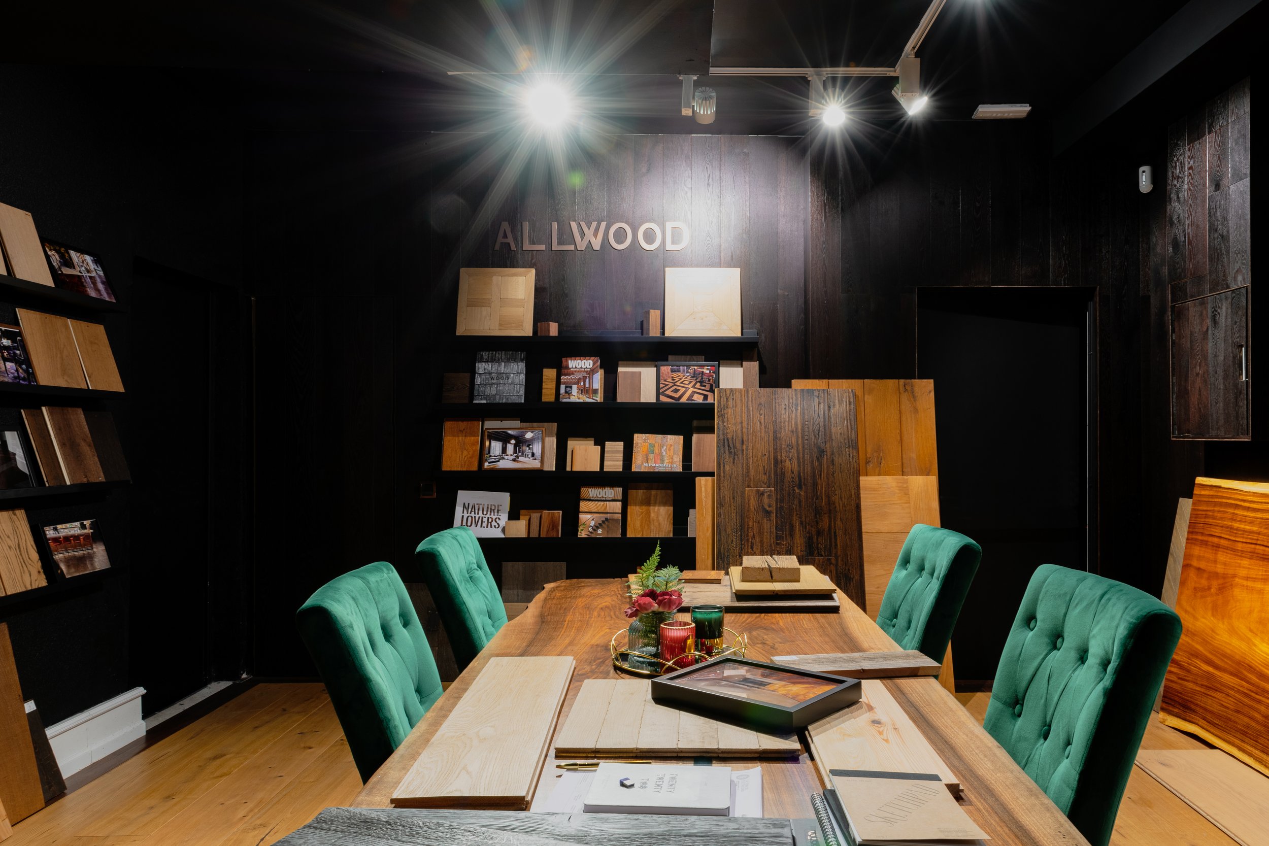Sala de exhibición de maderas y muestras, con una mesa y sillas verdes decorativas, en un espacio con paredes oscuras y estanterías con libros y láminas de madera, y un cartel que dice 'ALLWOOD'