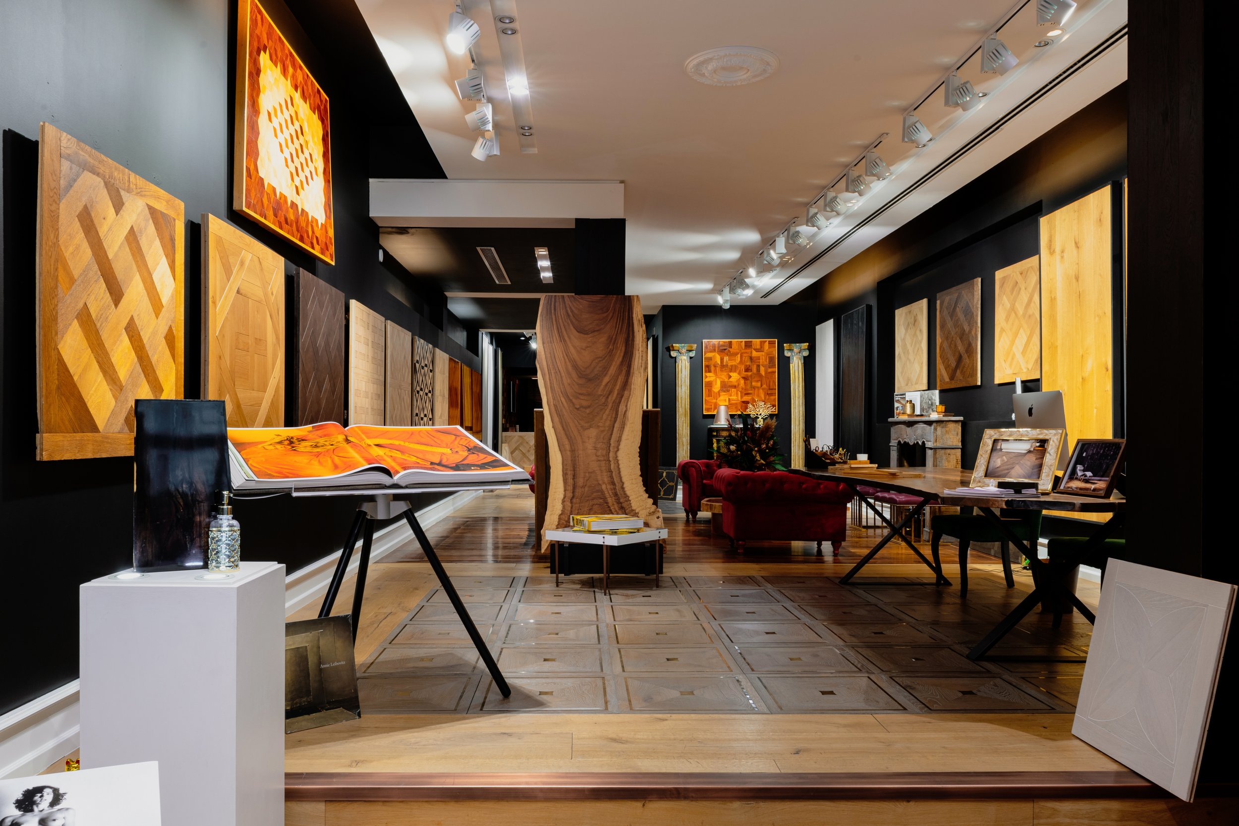 Sala de exhibición de obras de arte y madera decorativa, con cuadros, muebles y una mesa con libros.