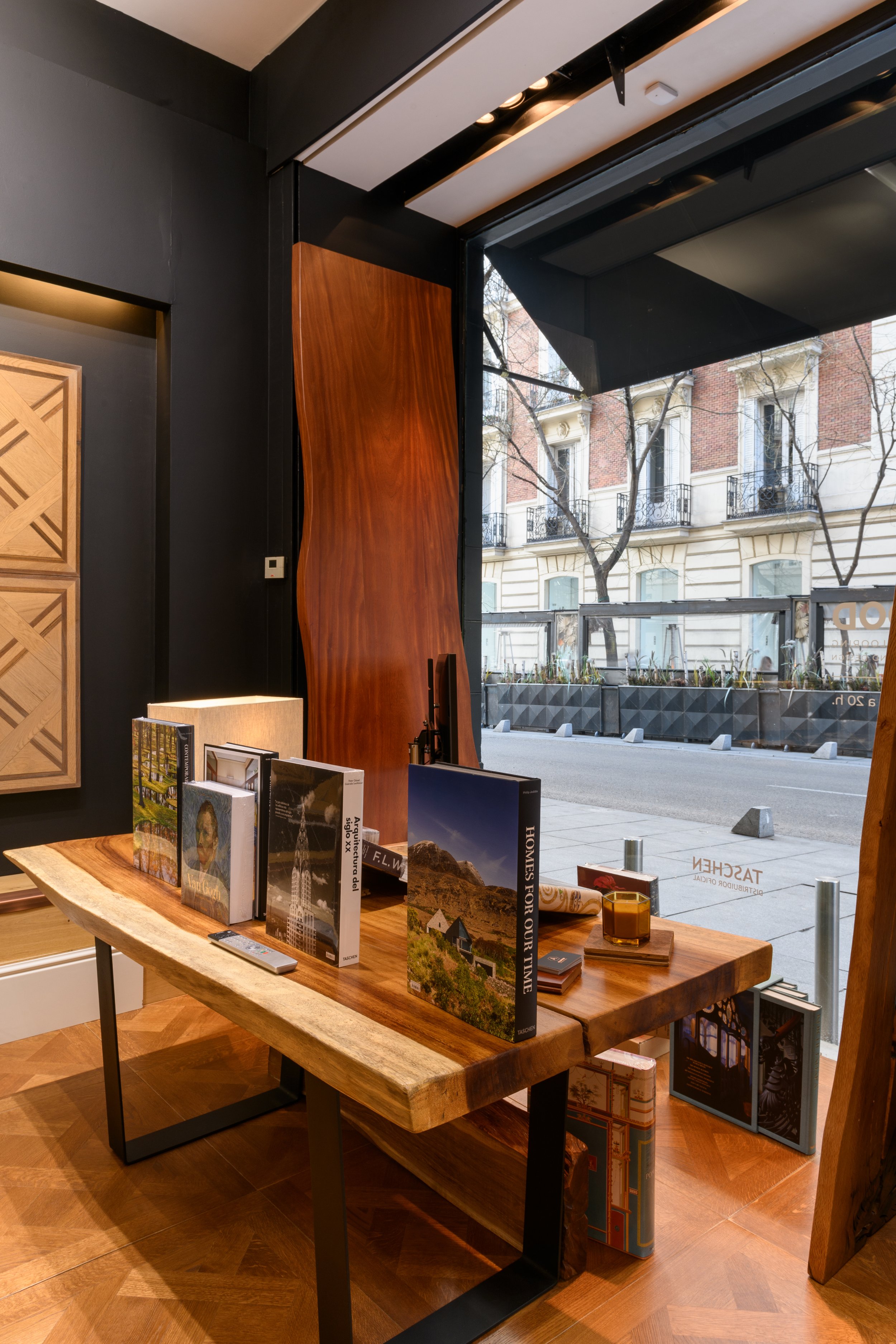 Vista interior de una librería con una mesa de madera con libros y una vela, junto a un ventanal con vista a la calle y edificios antiguos.