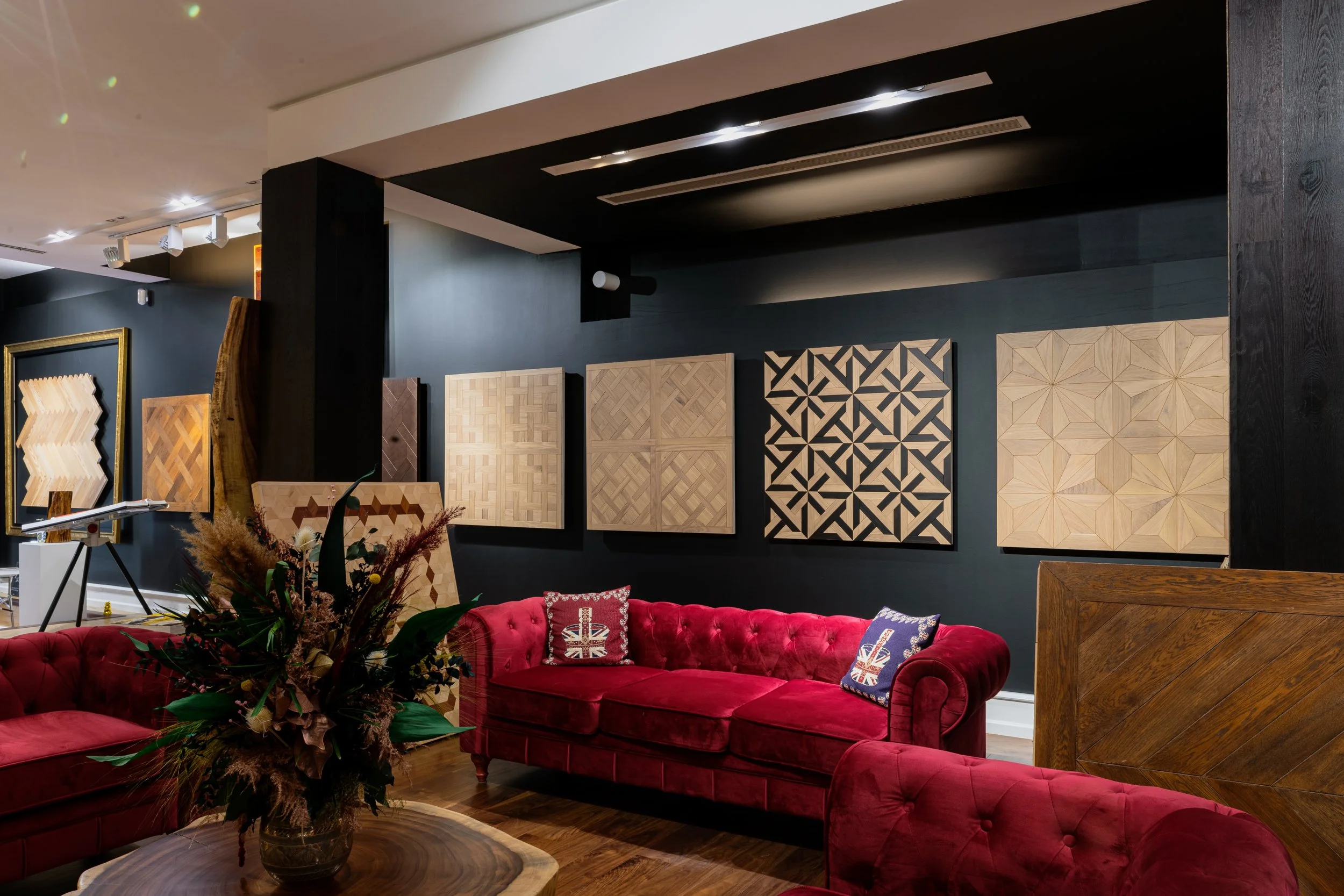 Sala de exposición con paredes negras, cuadros adornados con grabados en diferentes patrones de madera, y sillones de terciopelo rojo con cojines decorativos con motivos del Reino Unido.