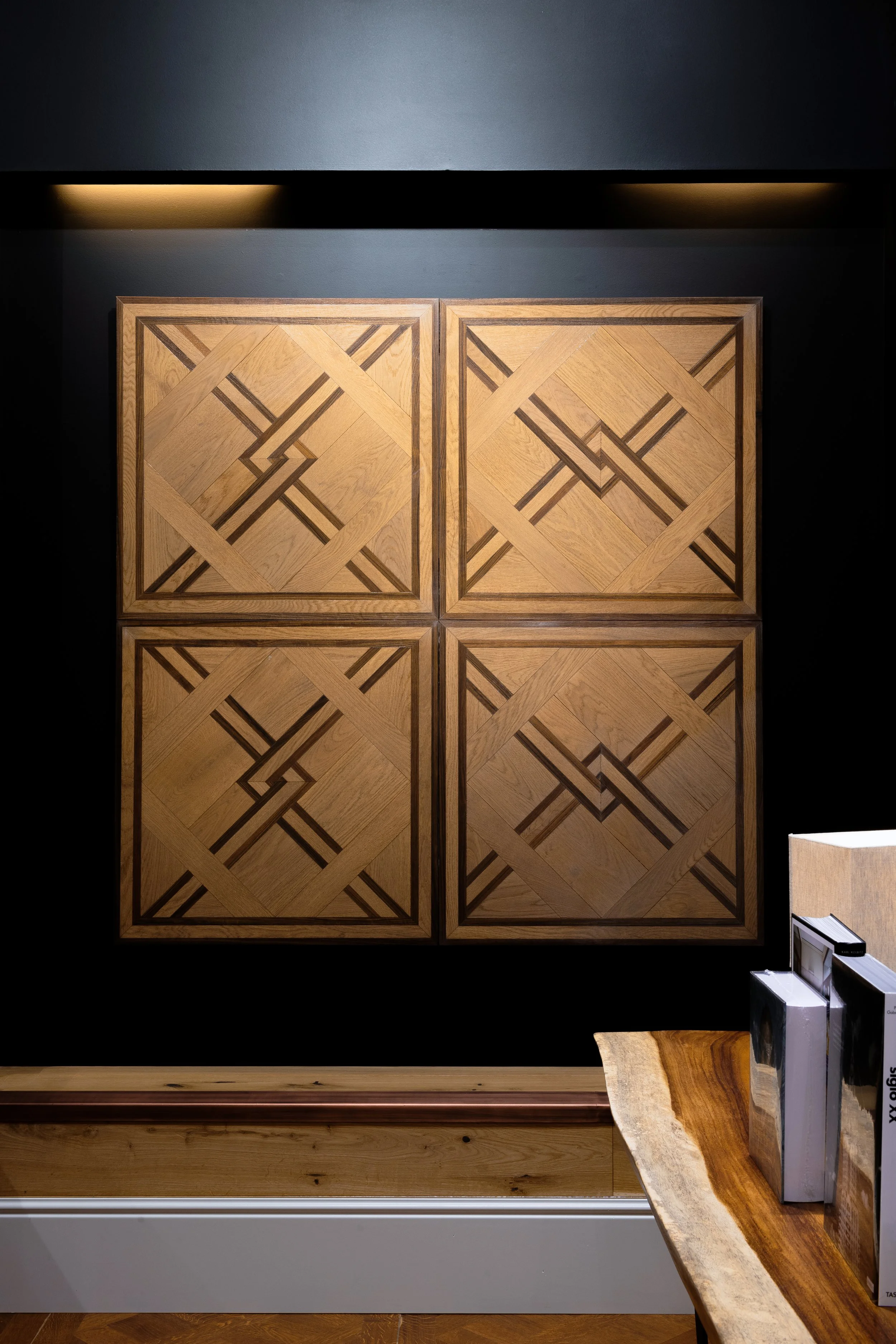 Cuatro paneles de madera con diseño geométrico en una pared negra, luz suave en la parte superior y mesa de madera con libros a la derecha.