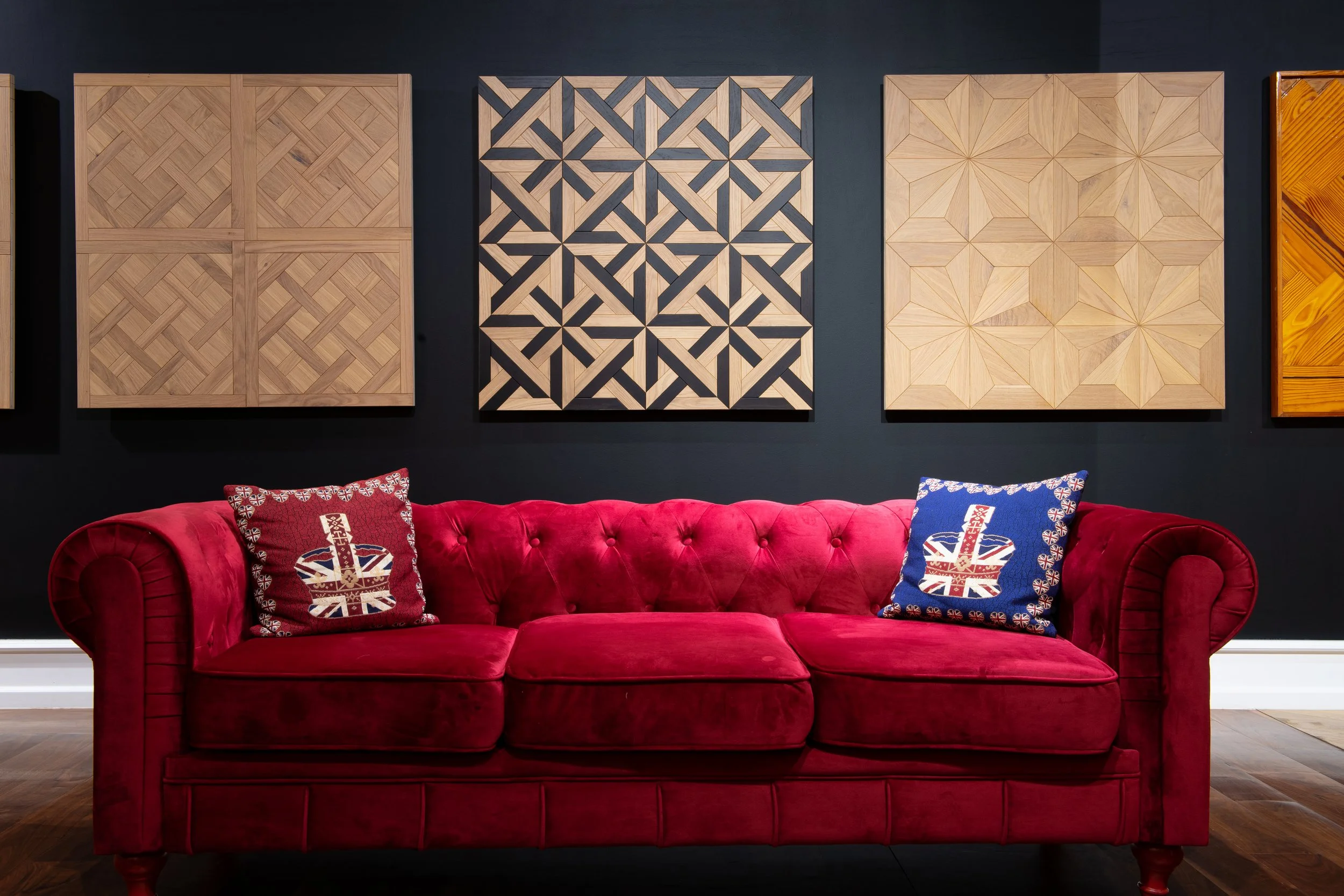 Sofá rojo de terciopelo con dos cojines que tienen banderas del Reino Unido, en una sala con paredes negras y obra de arte en madera en el fondo.