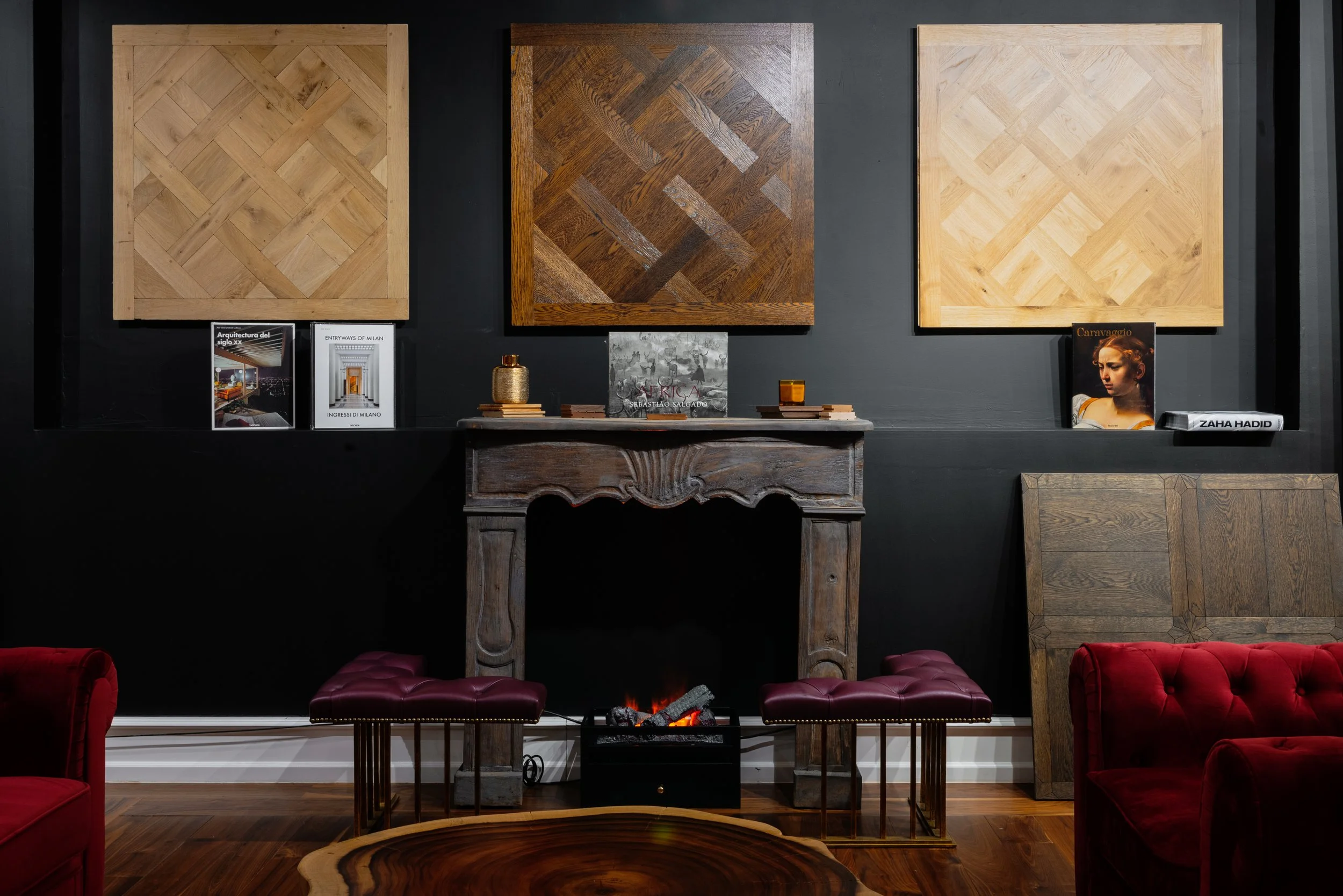 Sala de estar con paredes negras decoradas con tres cuadros de madera en diferentes tonos y patrones, un chimenea decorativa encendida, dos bancos con patas doradas y cojines de piel burdeos, un mueble que parece un tocador de madera vintage, y un so