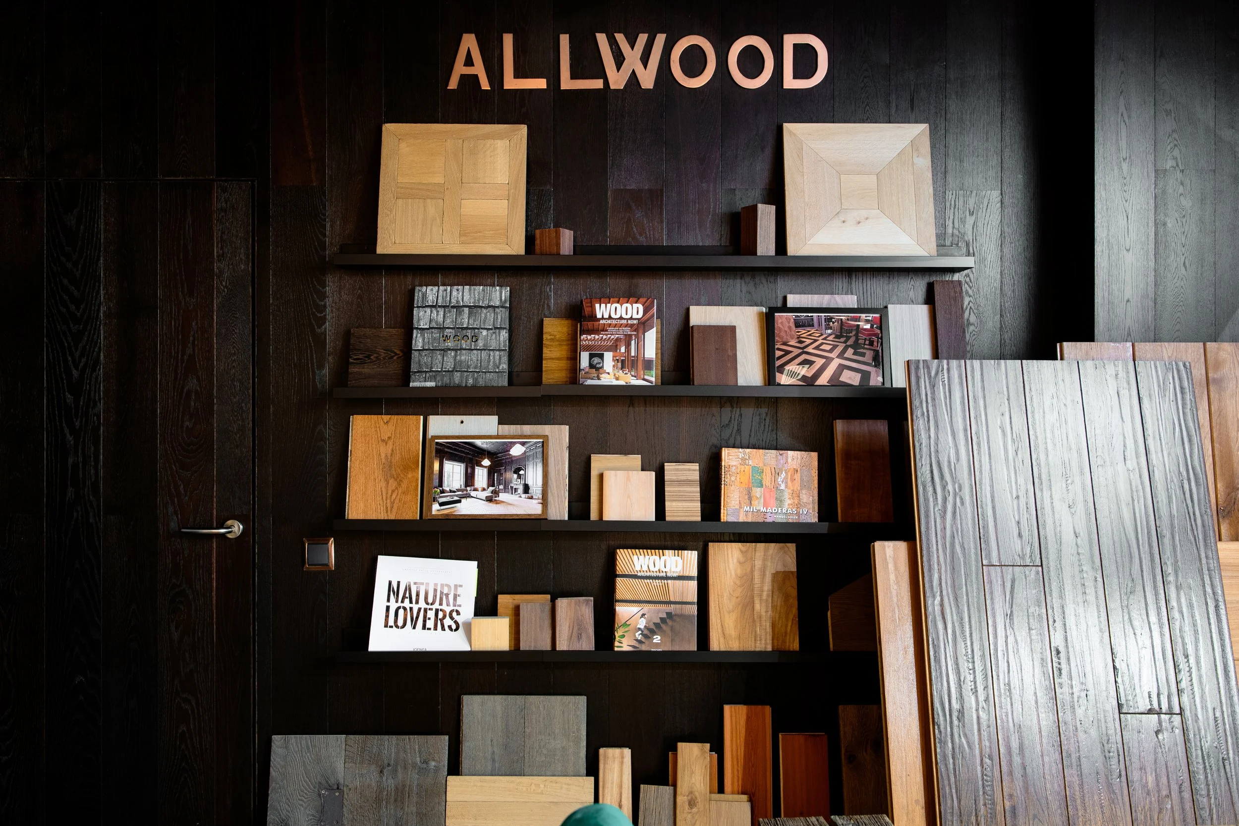 Estanterías de madera con muestras y catálogos de diseño de interiores contra una pared oscura con letras que dicen 'ALLWOOD' en la parte superior