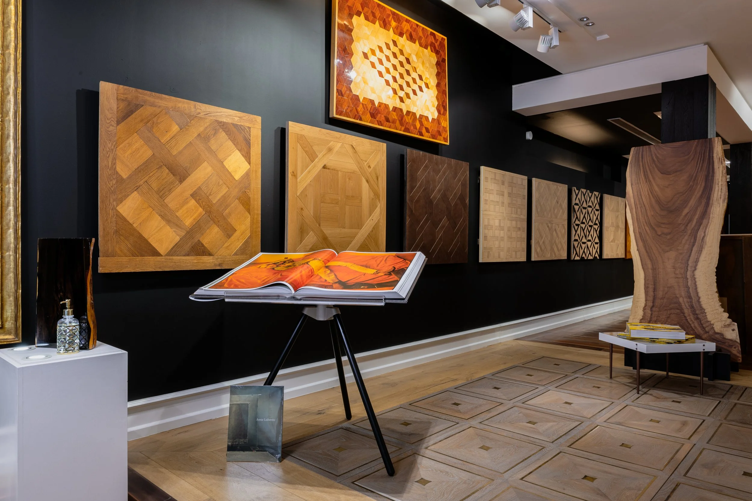 Galería de exhibición de tablas de madera con diferentes diseños en una pared negra, con un libro abierto en un soporte y una escultura de madera en un rincón