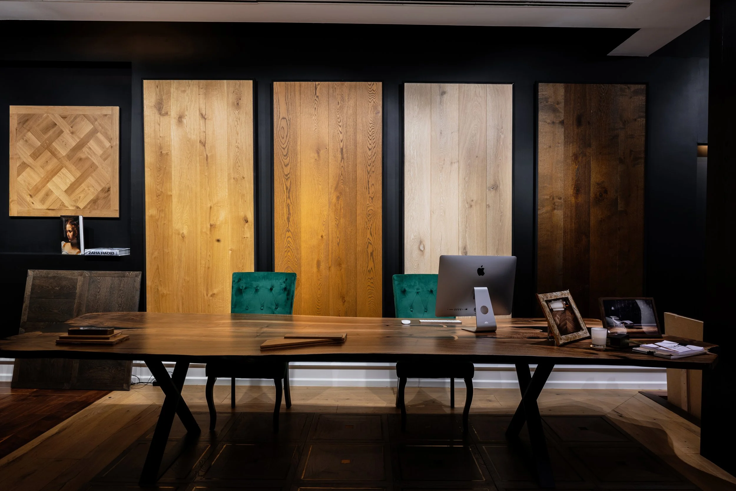 Una oficina con mesa de madera, una computadora iMac y sillas verdes. Fondo con paredes negras y paneles de madera de diferentes tonos.