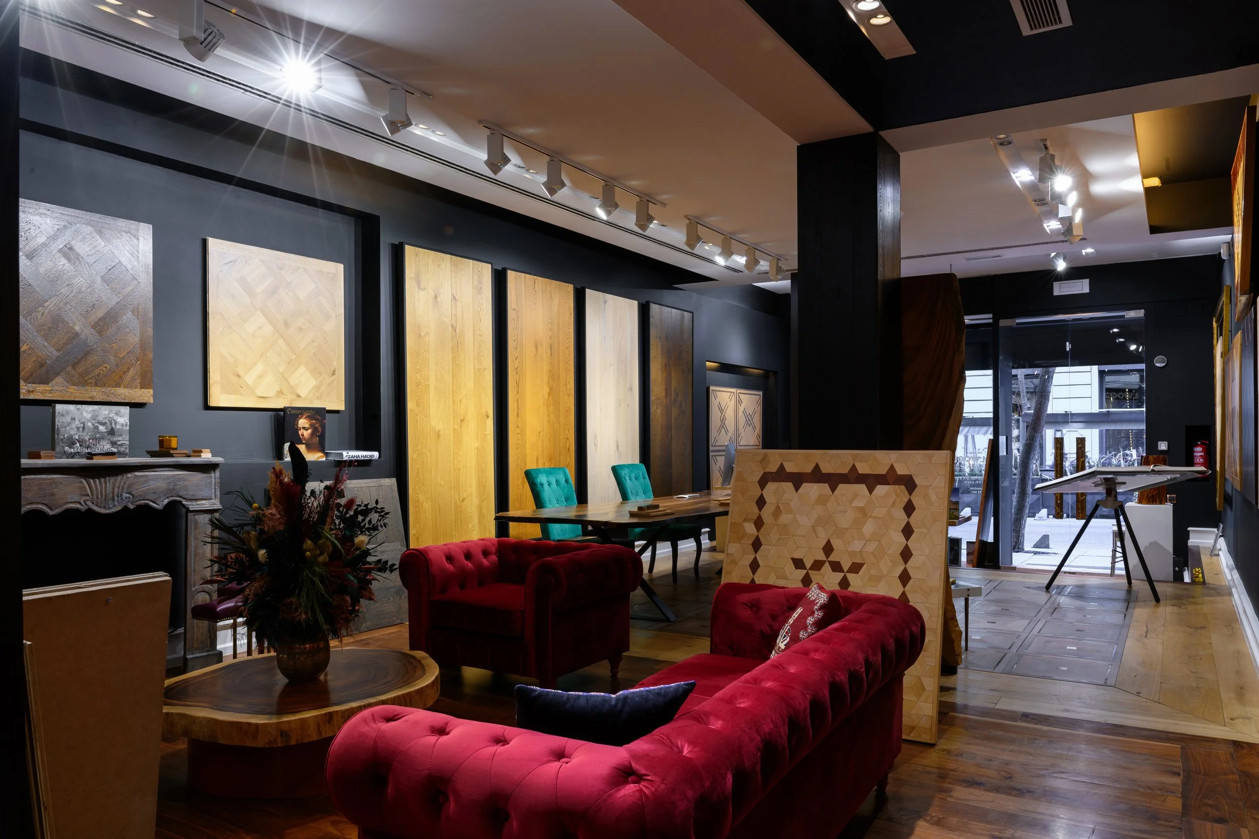 Sala de exposición con paredes de colores oscuros, cuadros de madera decorativos, muebles de sillones y mesas, y grandes puertas de vidrio al fondo con vista hacia la calle.