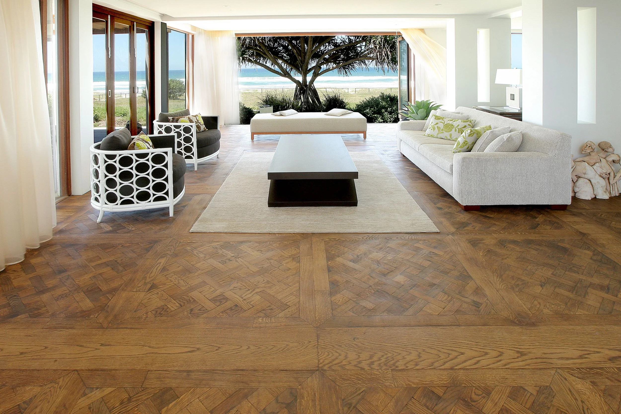 Sala de estar con vista al mar, con muebles blancos y negros, suelo de madera con patrón, grandes ventanas correderas abiertas que muestran arena y olas, árbol grande en el fondo, cojines decorativos en el sofá.