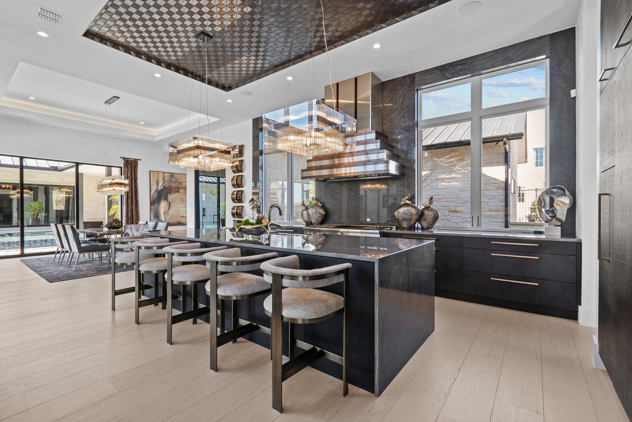 Interiores de una moderna cocina y comedor con luz natural, isla con sillas, lámparas decorativas, electrodomésticos y ventanas grandes.