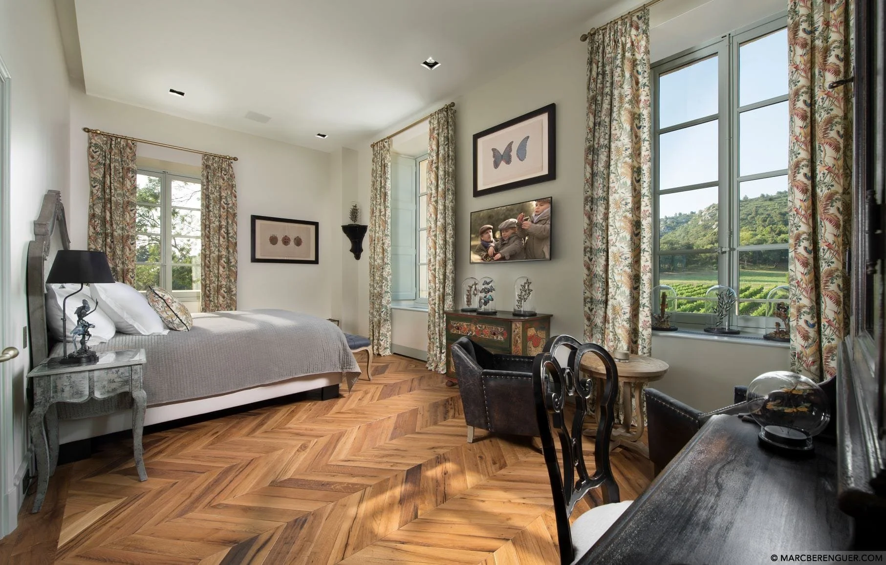 Habitación decorada con cortinas florales, cama con cabecera de madera, cuadros en las paredes, ventanas grandes con vista a un campo y muebles vintage.