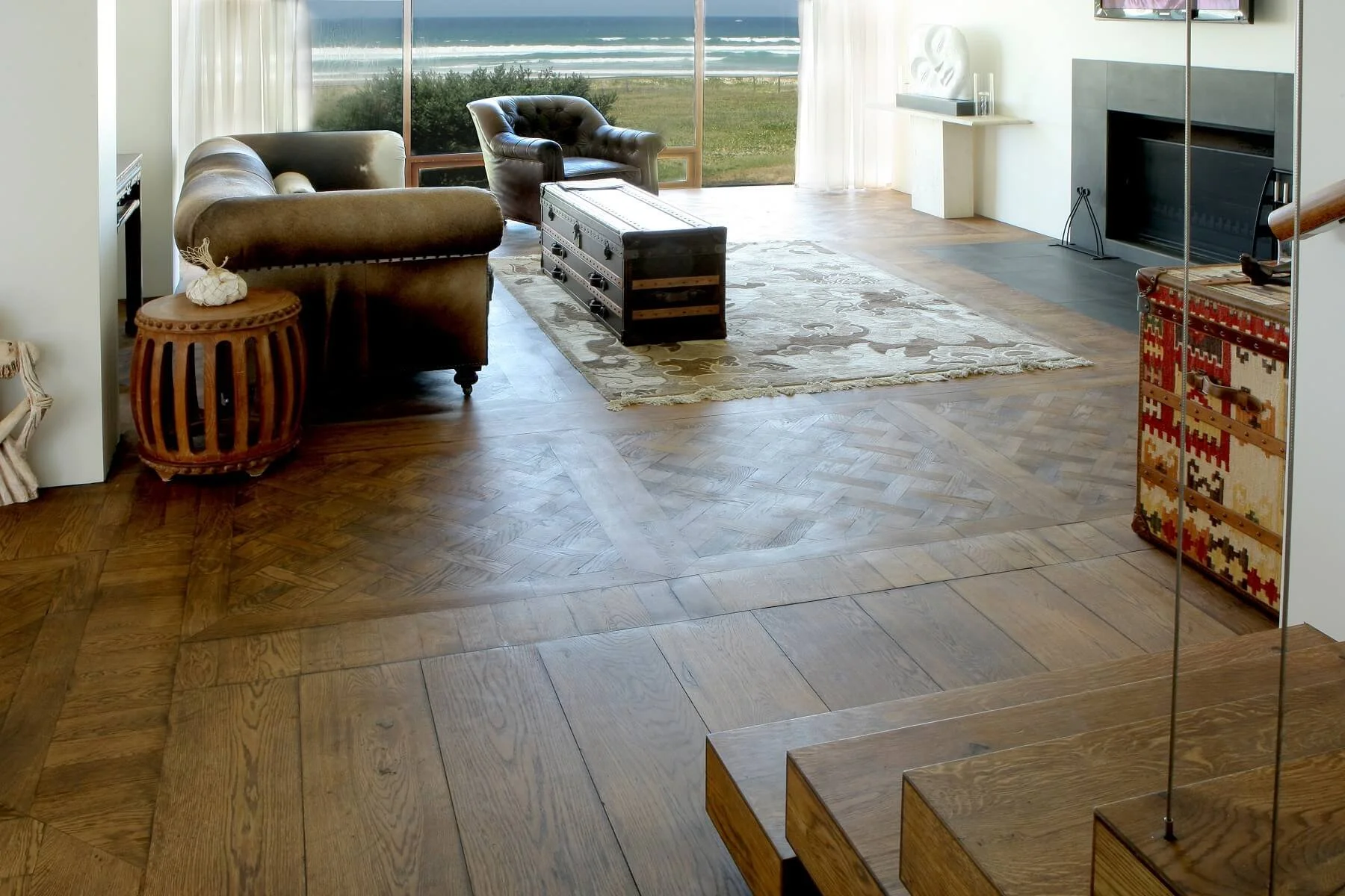 Sala de estar con sillones de cuero, una alfombra y una vista al exterior a través de ventanas grandes con cortinas blancas.