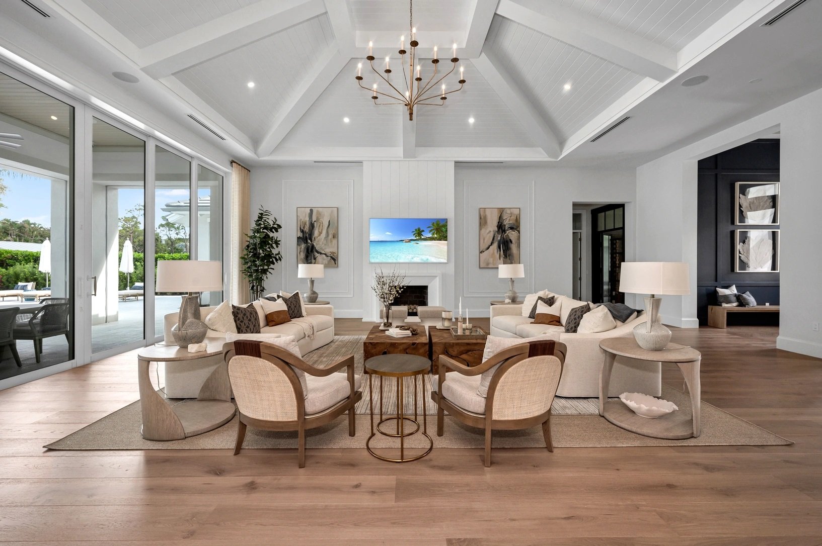 suelos de madera en sala de estar moderna con sillones claros, mesas de centro de madera y lámparas. Pared con arte y televisor de paisaje en la pared de fondo. Grandes puertas de vidrio que muestran un patio con sombrillas y sillas.