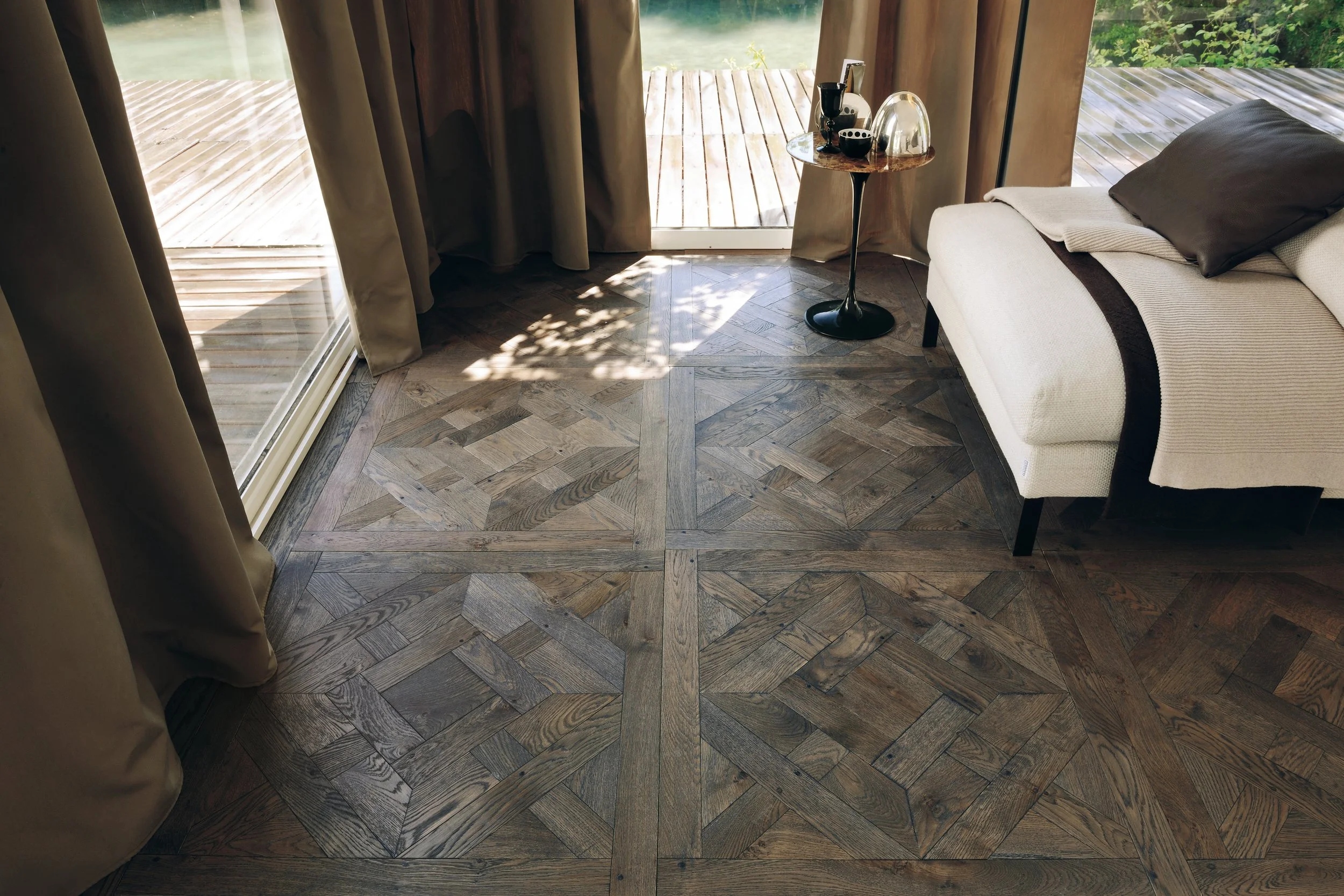 Interior de sala con piso de madera en patrón geométrico, cortinas beige, sillón blanco, mesa pequeña con bebidas y objetos decorativos, vista a un deck de madera y áreas verdes exteriores.