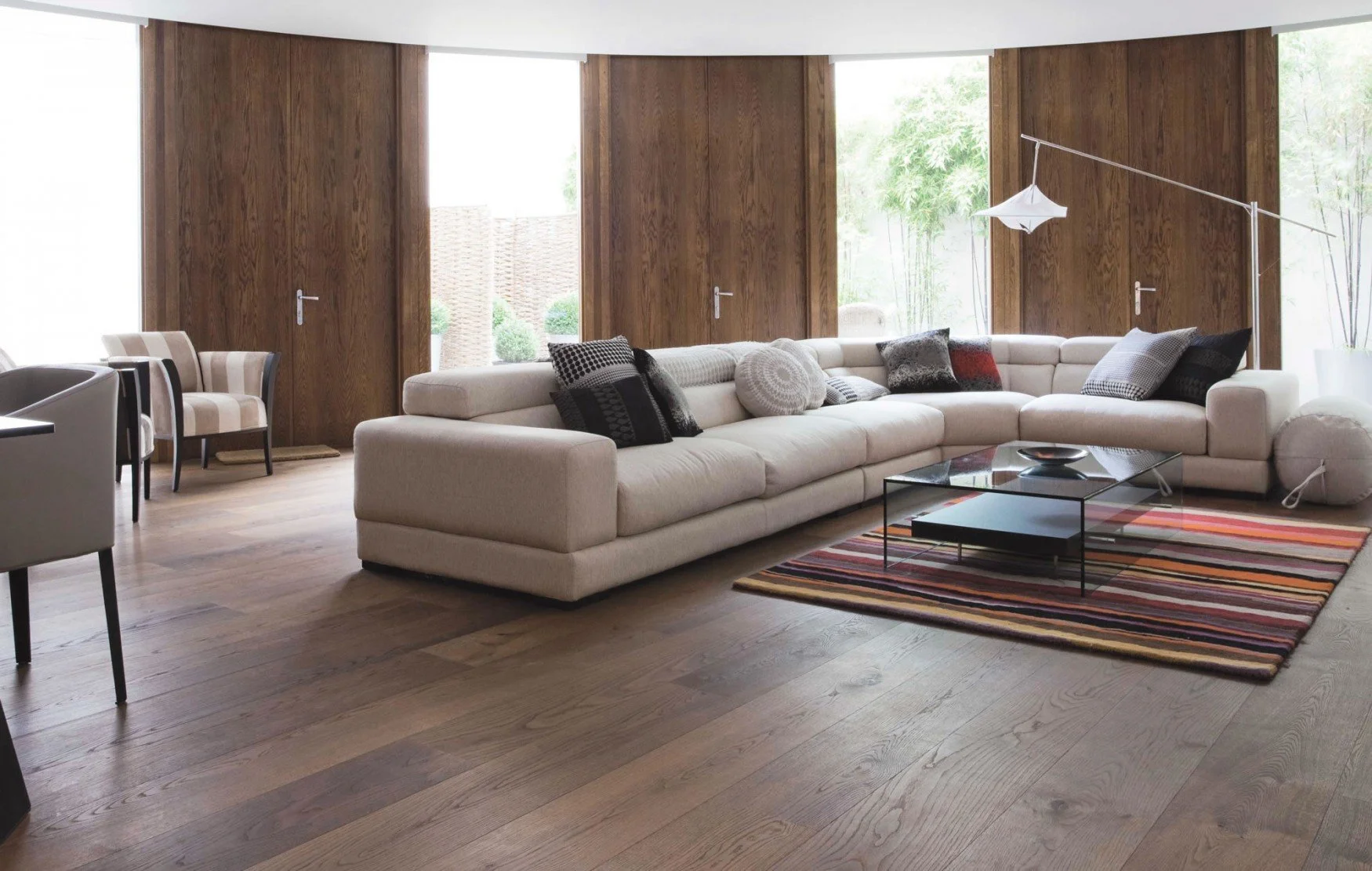 Sala de estar con sofá modular grande en tonos claros, varias almohadas decorativas en colores oscuros, mesa de centro de vidrio con marco negro, alfombra de rayas en tonos terracota, madera en las paredes y piso, con ventanas grandes que dejan pasar