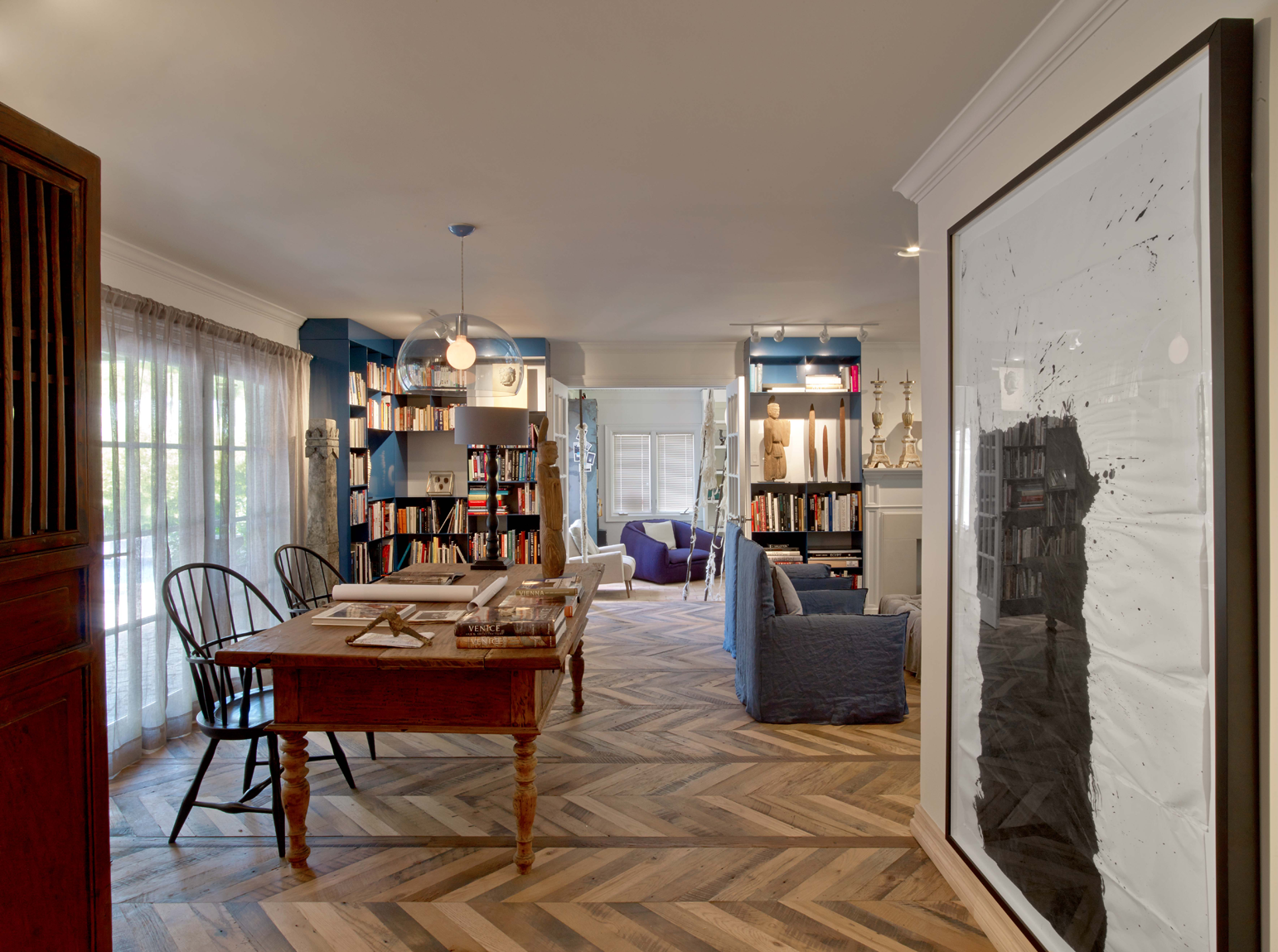 Sala de estar con diván azul, estanterías llenas de libros y decoración en tonos artísticos, pisos de madera y grandes ventanas con cortinas blancas.