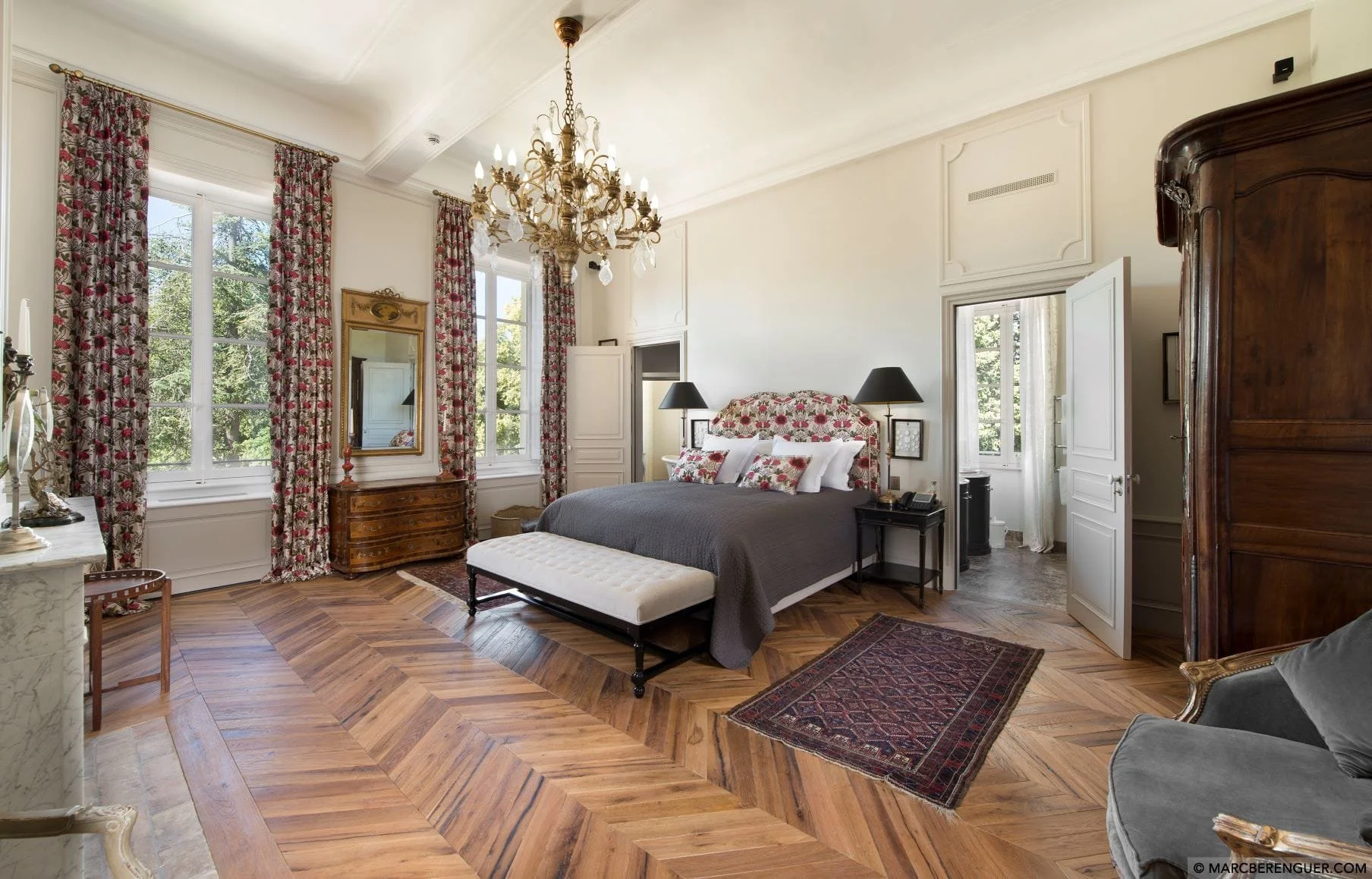 Habitación elegante con ventanas grandes, cortinas con flores, cama con cabecera floral, lámparas de noche negras, alfombra antigua y muebles de madera.
