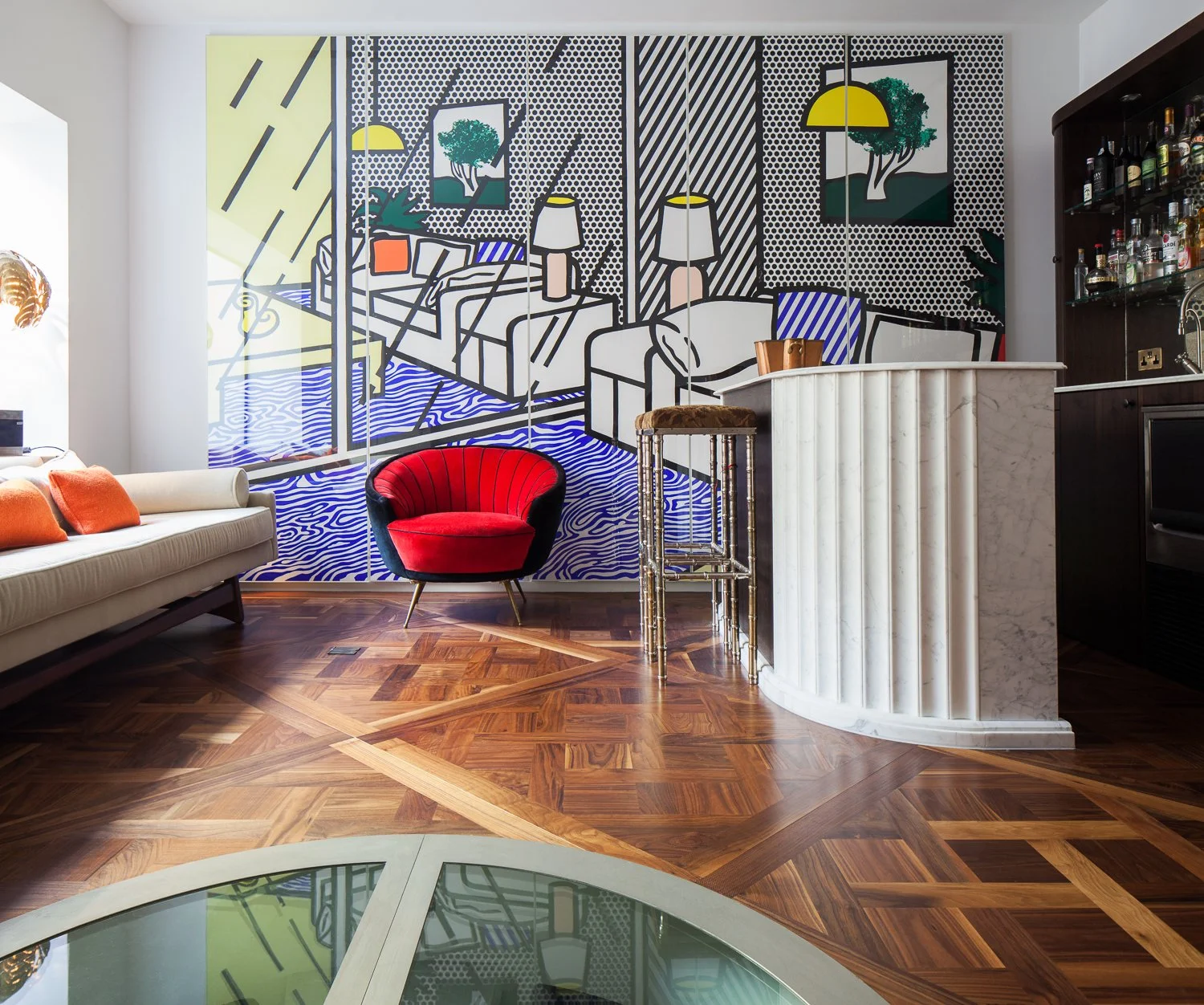 Sala de estar moderna con pared decorativa de arte en estilo pop art, sillón rojo y sofá beige con cojines anaranjados, suelo de madera con patrón, barra de mármol y estantería de bebidas.