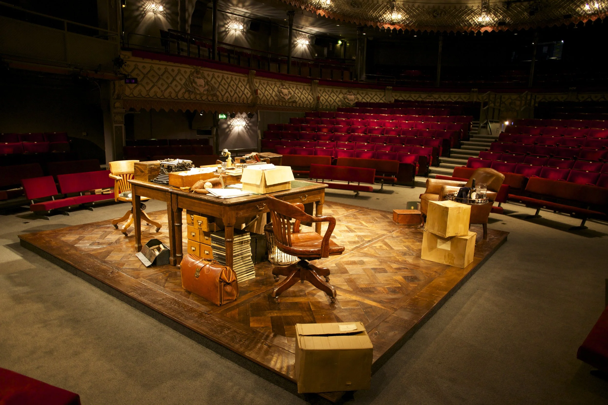 Escenario teatral con escritorio lleno de papeles, libros, y objetos, rodeado por sillas tapizadas de color burdeos para audiencia, en un teatro vacío.