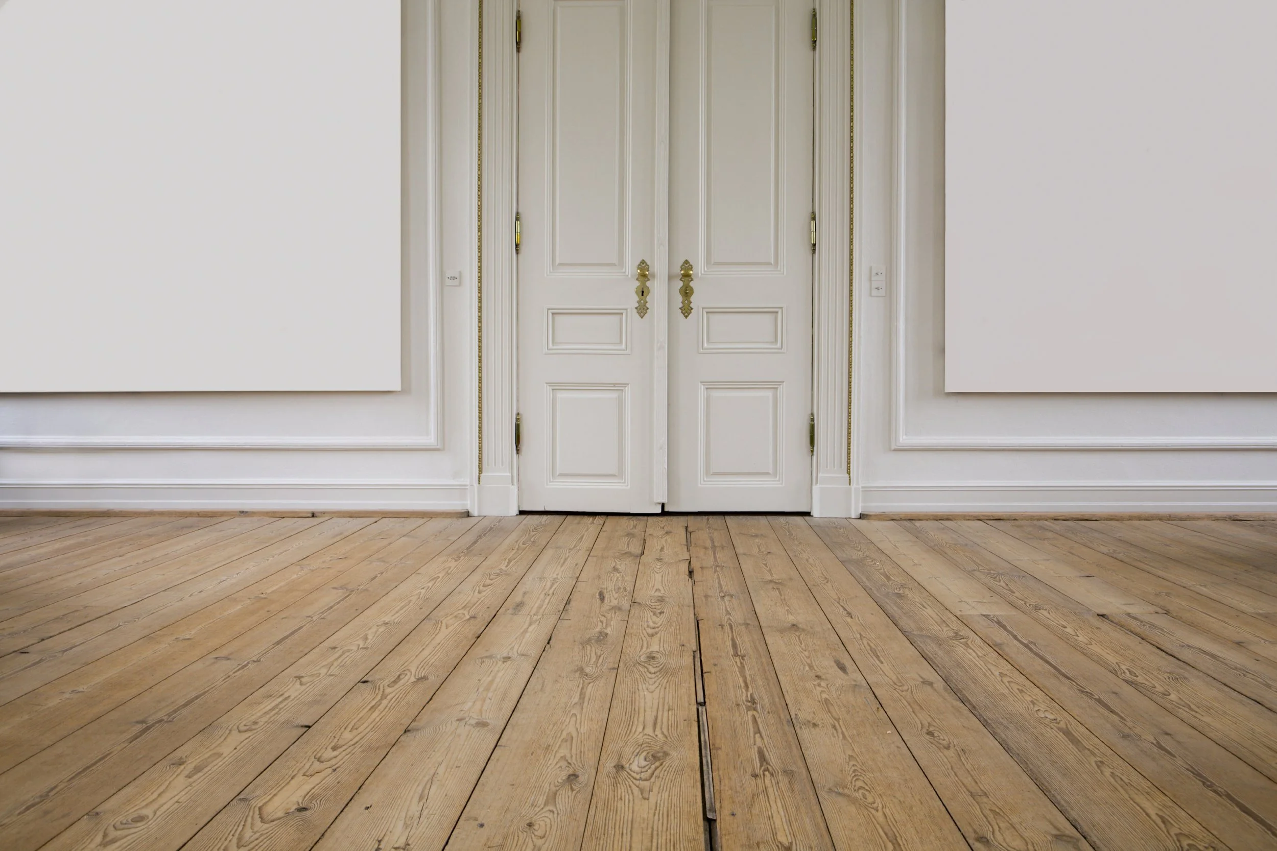 Puerta doble blanca con detalles dorados en un pared blanca con piso de madera.