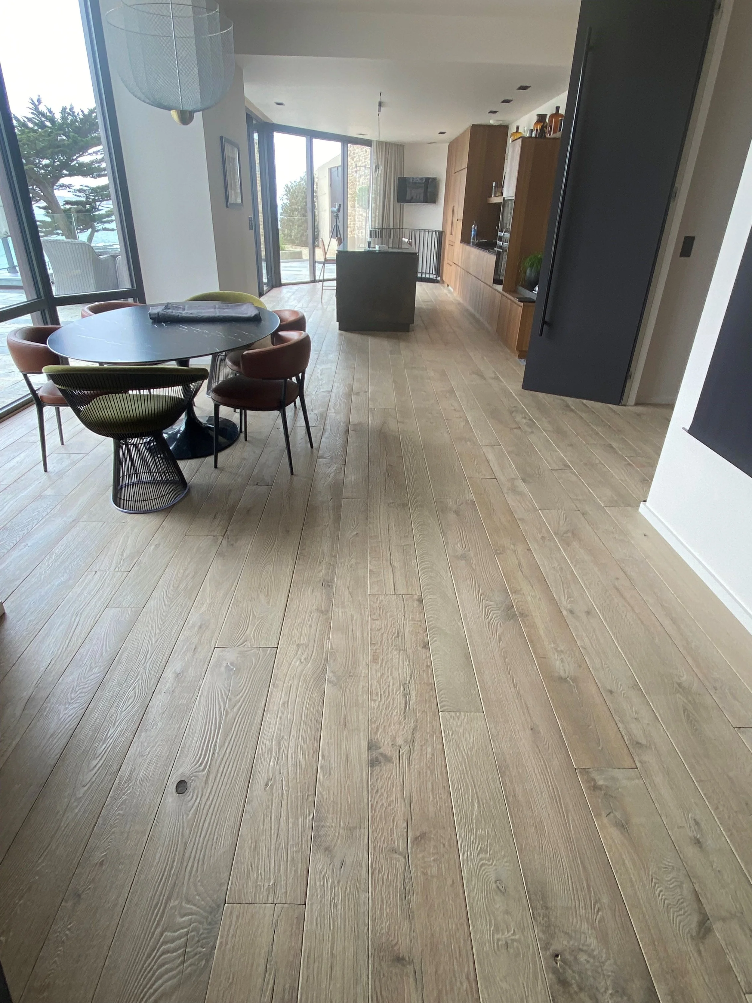 Interior de una sala de comedor y cocina en una casa moderna con pisos de madera, mesa redonda con sillas alrededor y ventanas grandes con vista al exterior