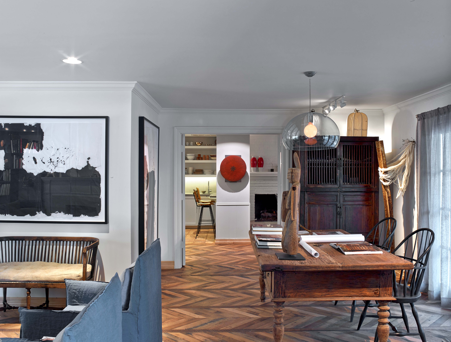 Sala de estar con pared blanca y piso de madera en espiga. Un sillón de madera con cojines y un sofá azul. Cuadros en la pared. Mesa de comedor de madera con objetos decorativos y libros, alrededor de ella cuatro sillas negras. En el fondo, una pared