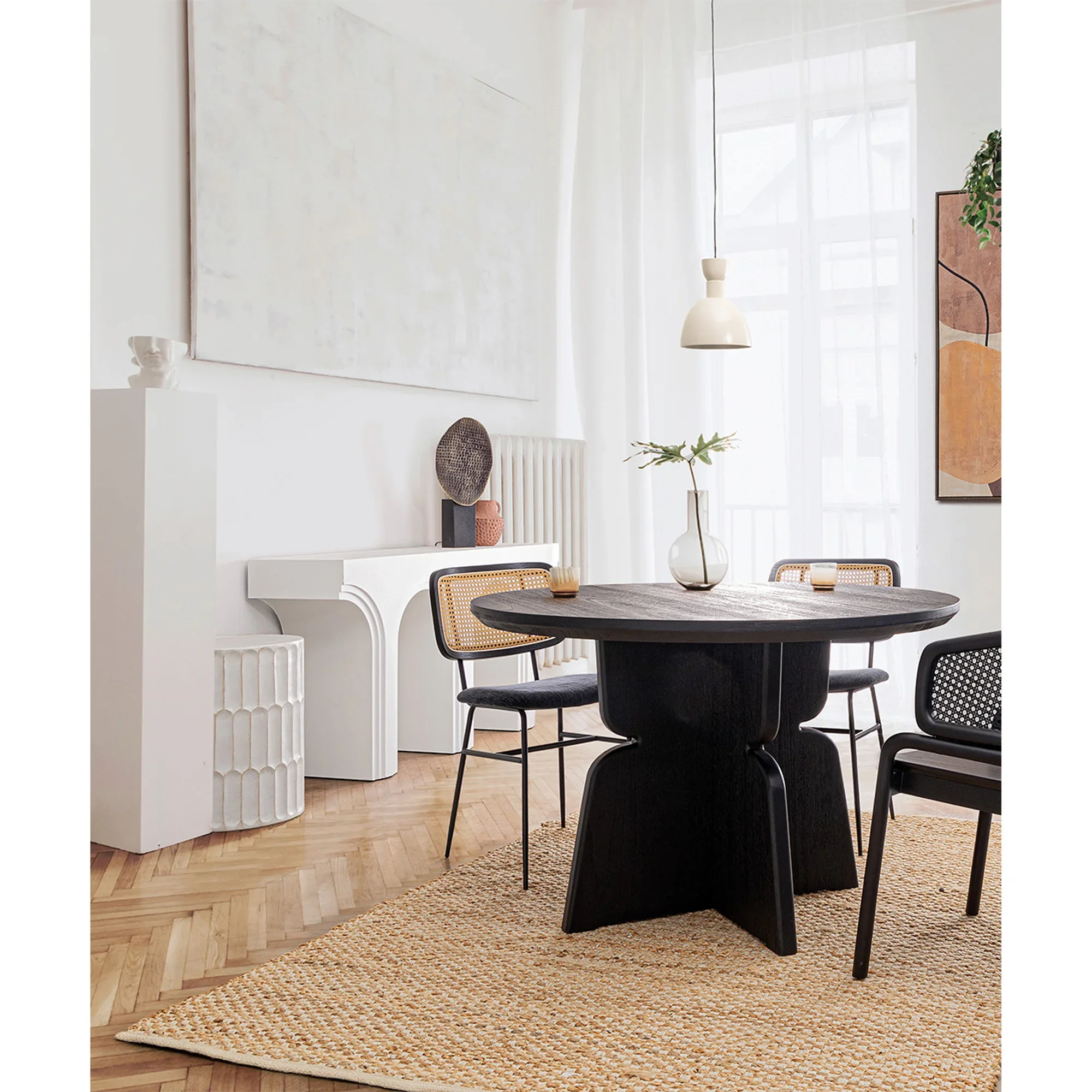 Sala de comedor con mesa redonda de madera oscura, sillas negras con respaldo de mimbre, alfombra de fibra, cortinas blancas, paredes blancas, decoraciones minimalistas y luz natural.
