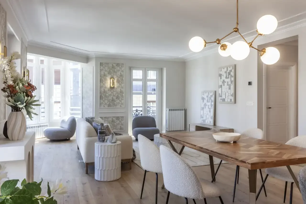 Sala de estar luminosa con sofá blanco, sillas y mesa de comedor de madera, flores en jarrón, paredes decoradas y ventanas grandes