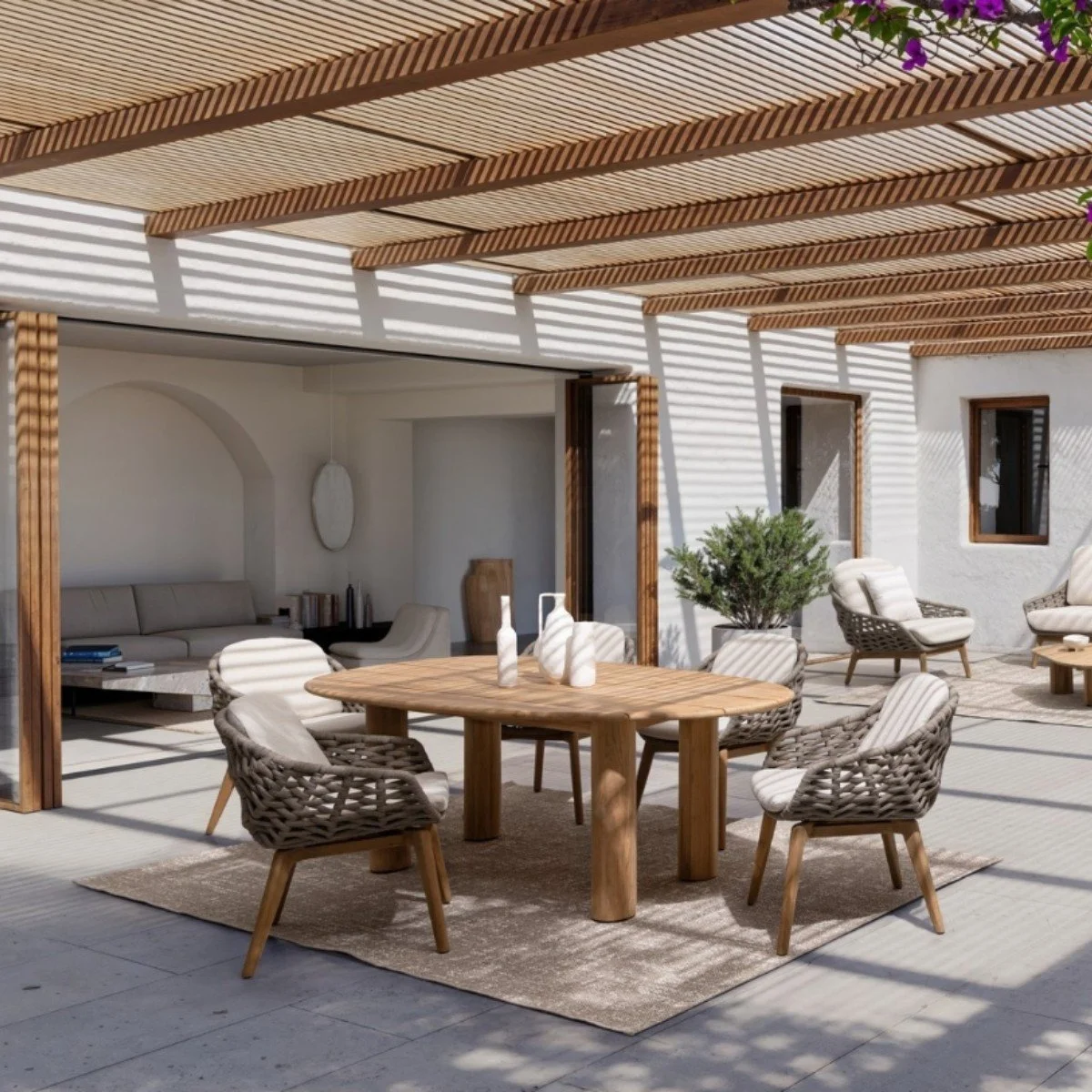 Terraza con mobiliario de madera, sillas con cojines, mesa de comedor, y sombra de listones de madera en el techo, en un ambiente exterior con paredes blancas y plantas decorativas.