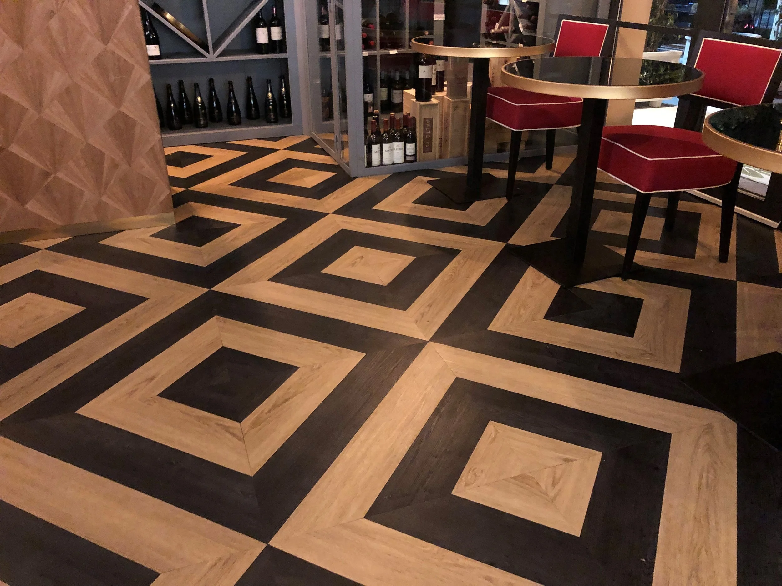 Interiores de un restaurante con mesas redondas con marcos dorados, sillas con cojines rojos y estantería con botellas de vino.