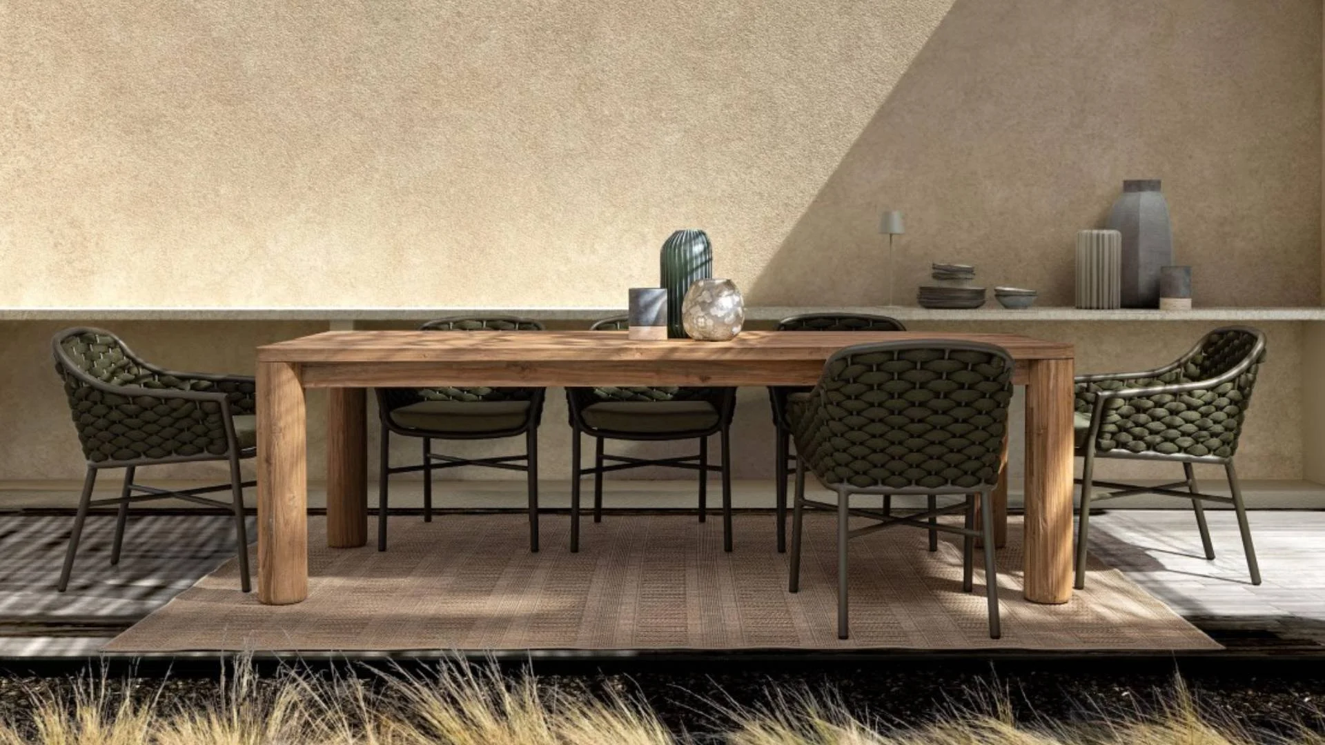 Mesa de madera con ocho sillas de estilo moderno, decorada con jarrones y objetos en una base de alfombra sobre el suelo y una pared beige con estantes decorativos.