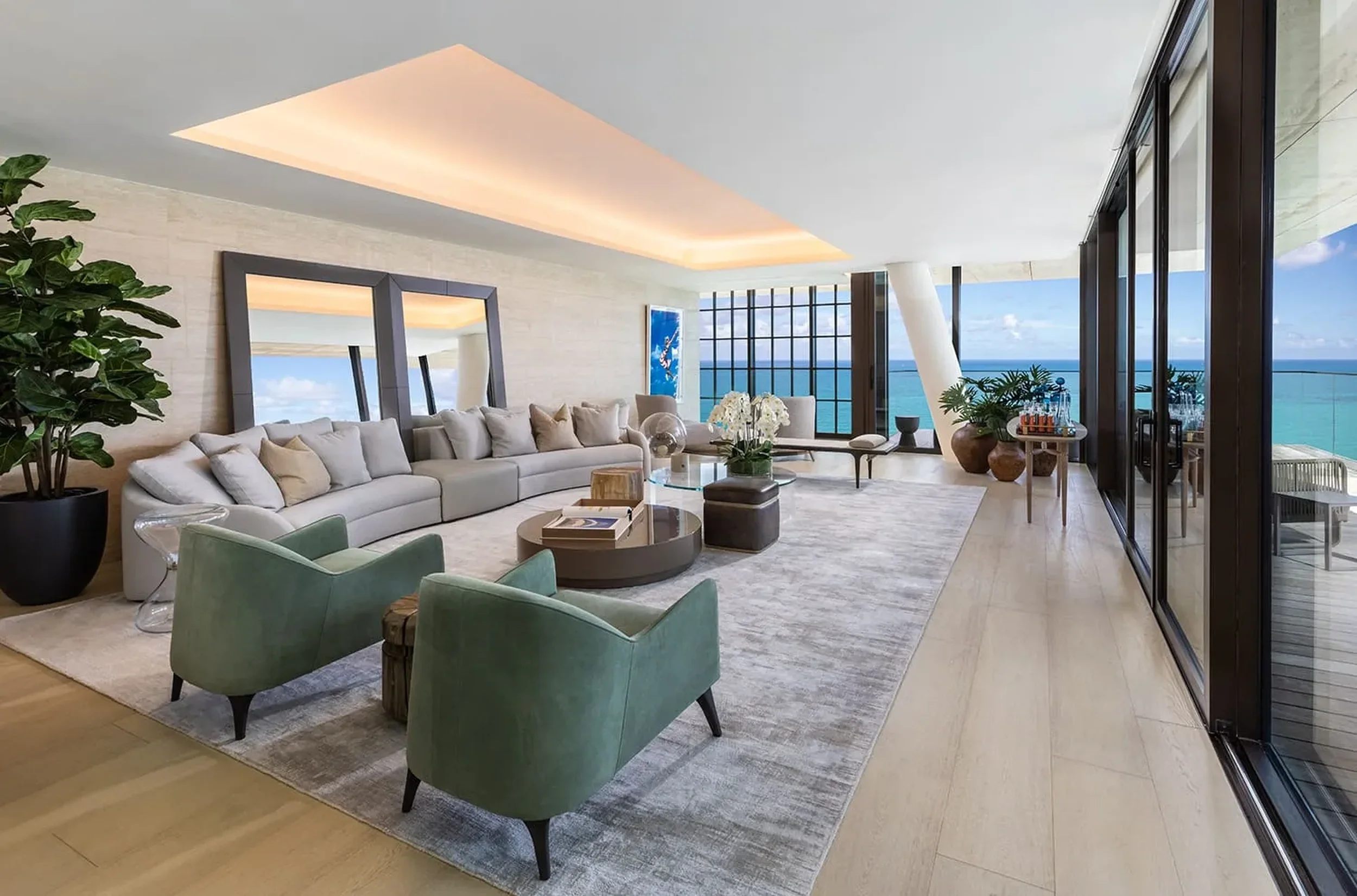 Sala moderna con vista al mar, sofá largo gris, sillones verdes, cuadro de arte en la pared, plantas, mesas de centro, lámpara de techo empotrada y grandes ventanales con vista al océano.