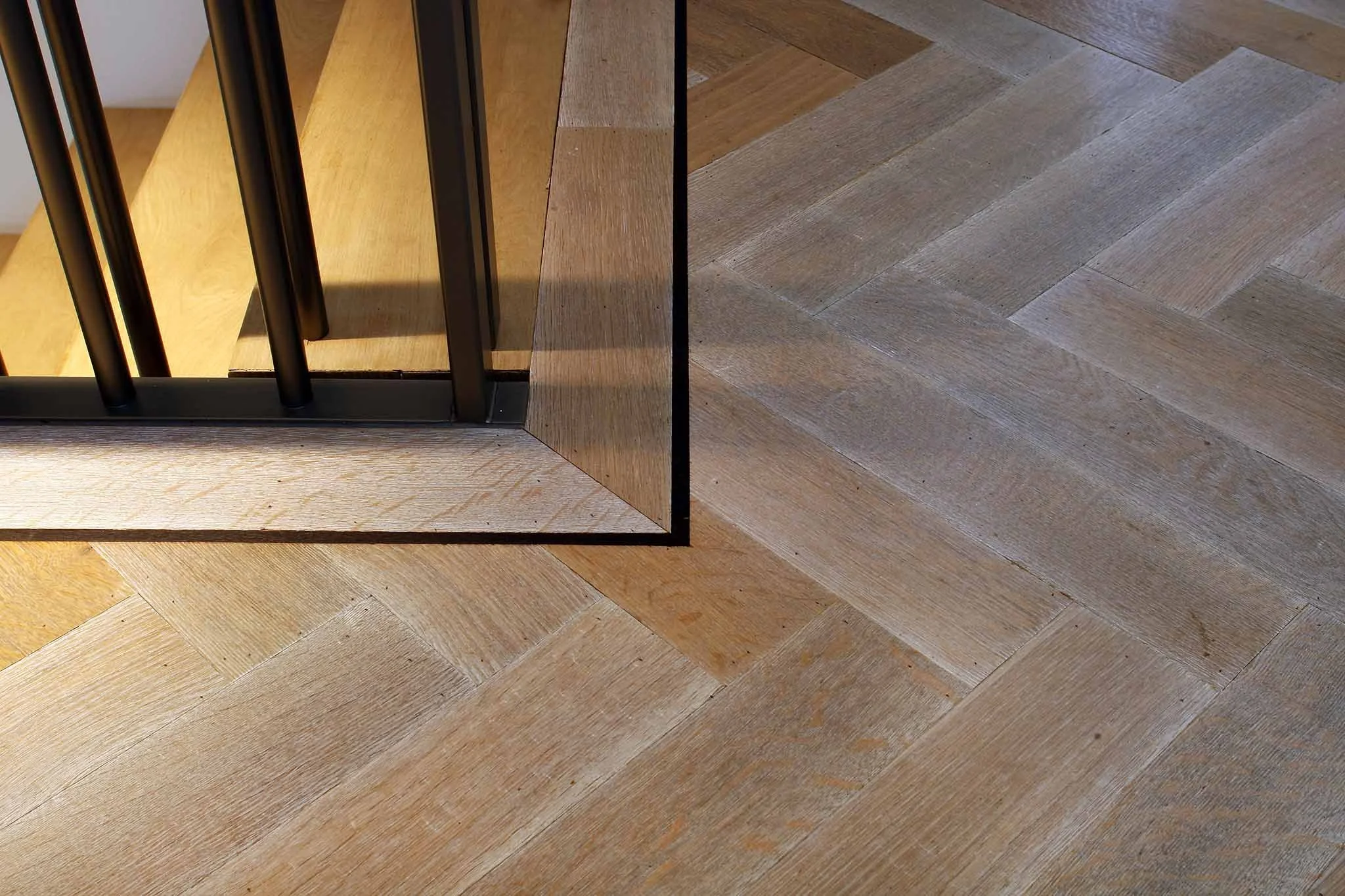 Vista de una esquina con suelo de madera en patrón ancho y un borde de madera clara, y una estructura metálica negra con barras verticales y panel de madera en el interior.