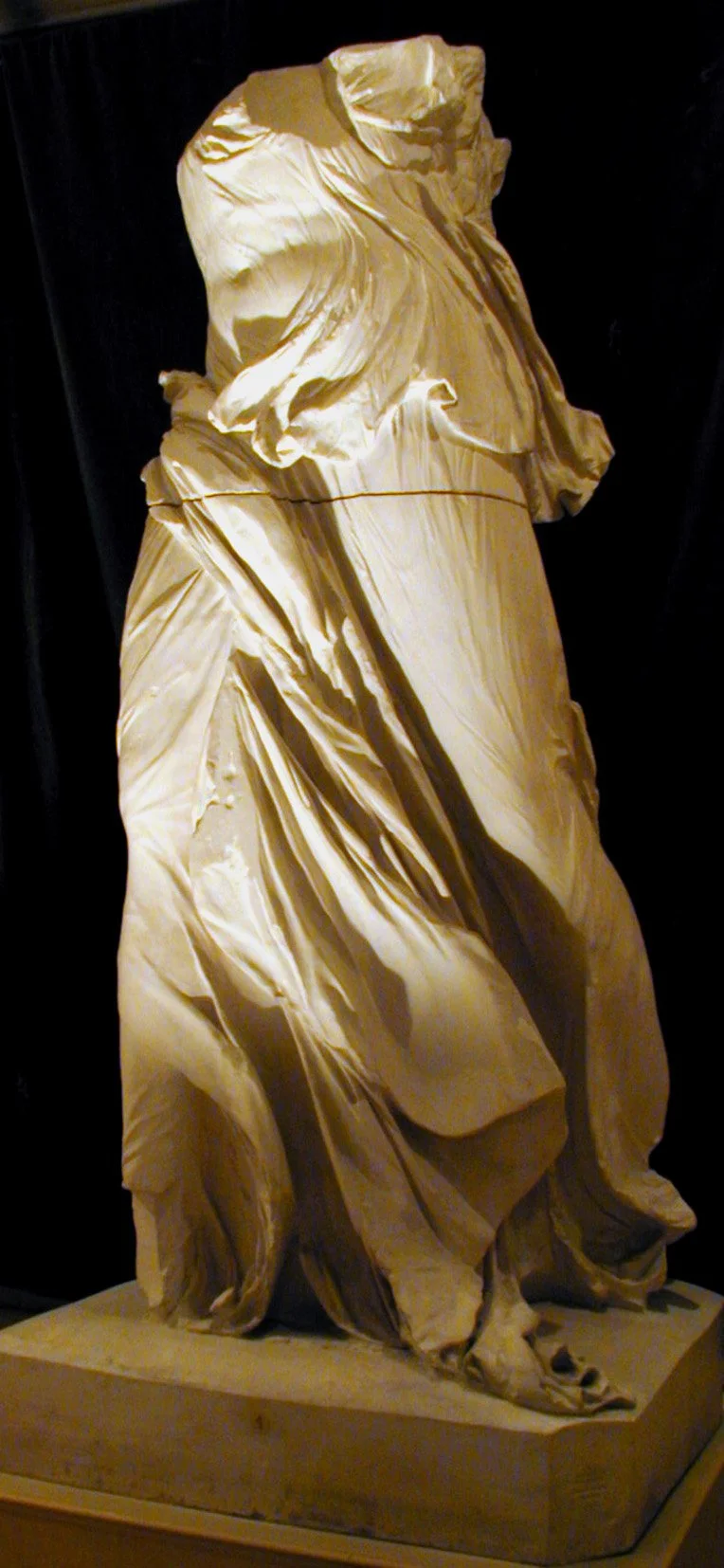 Escultura en mármol de una figura humana con ropajes ondulados y detalles en pliegues, exhibida en un fondo oscuro.