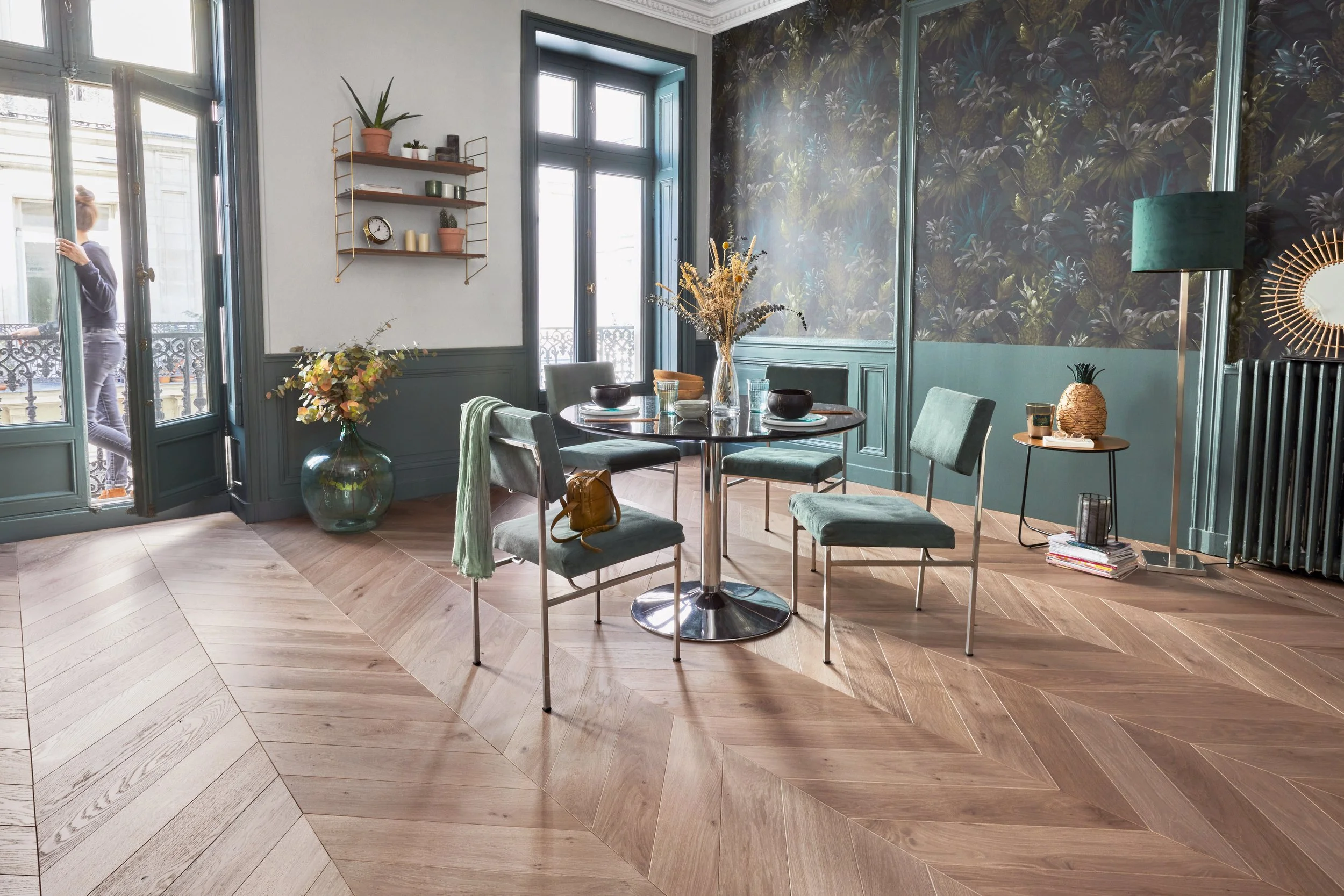 Sala de comedor con paredes decoradas con papel tapiz de hojas verdes, varios muebles y plantas, una mesa redonda con sillas de tela azul y decoraciones.