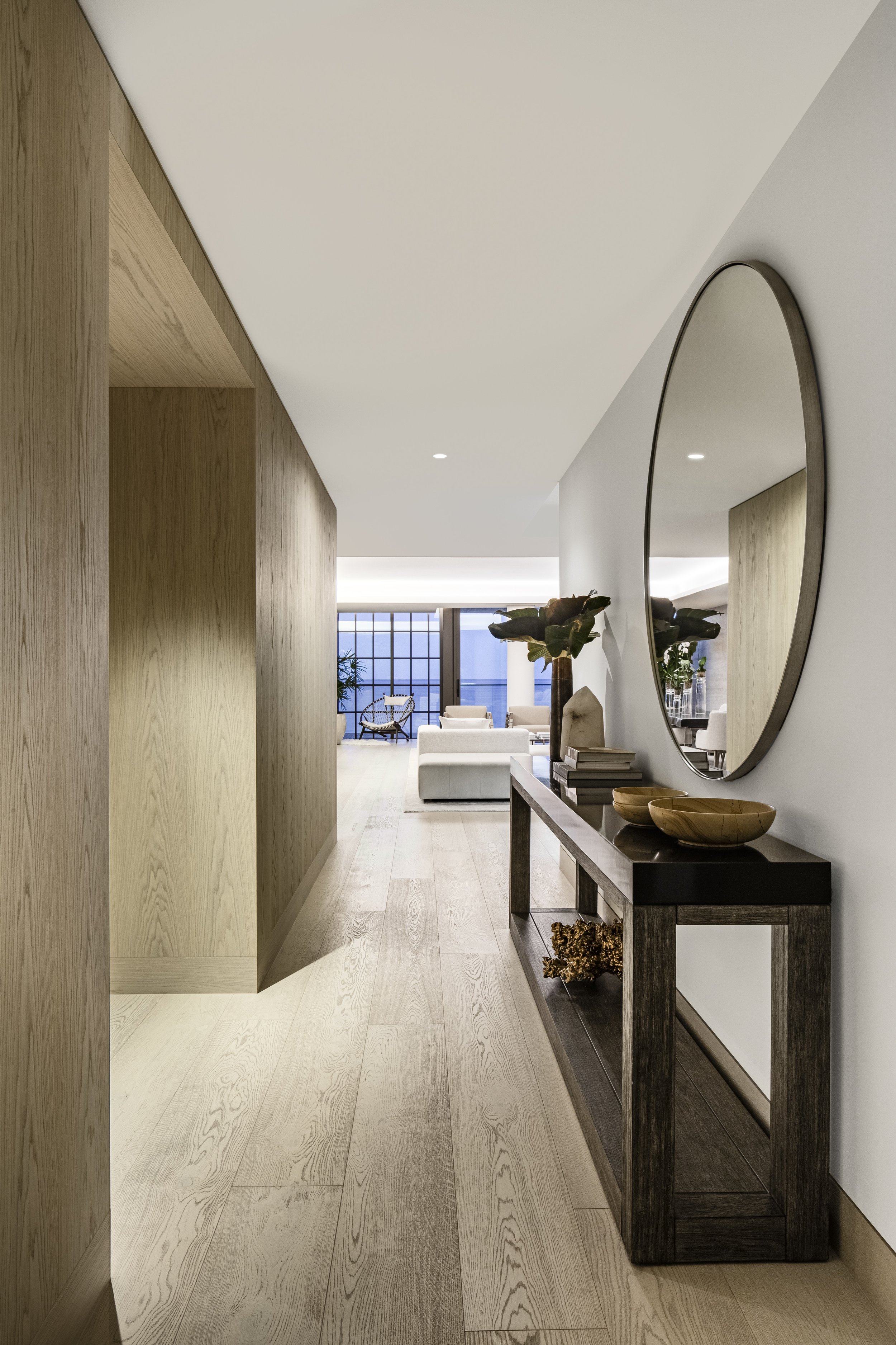 suelos de madera en pasillo con mesa de madera, espejo redondo en la pared y vista a una sala con sillones blancos y ventanas grandes.