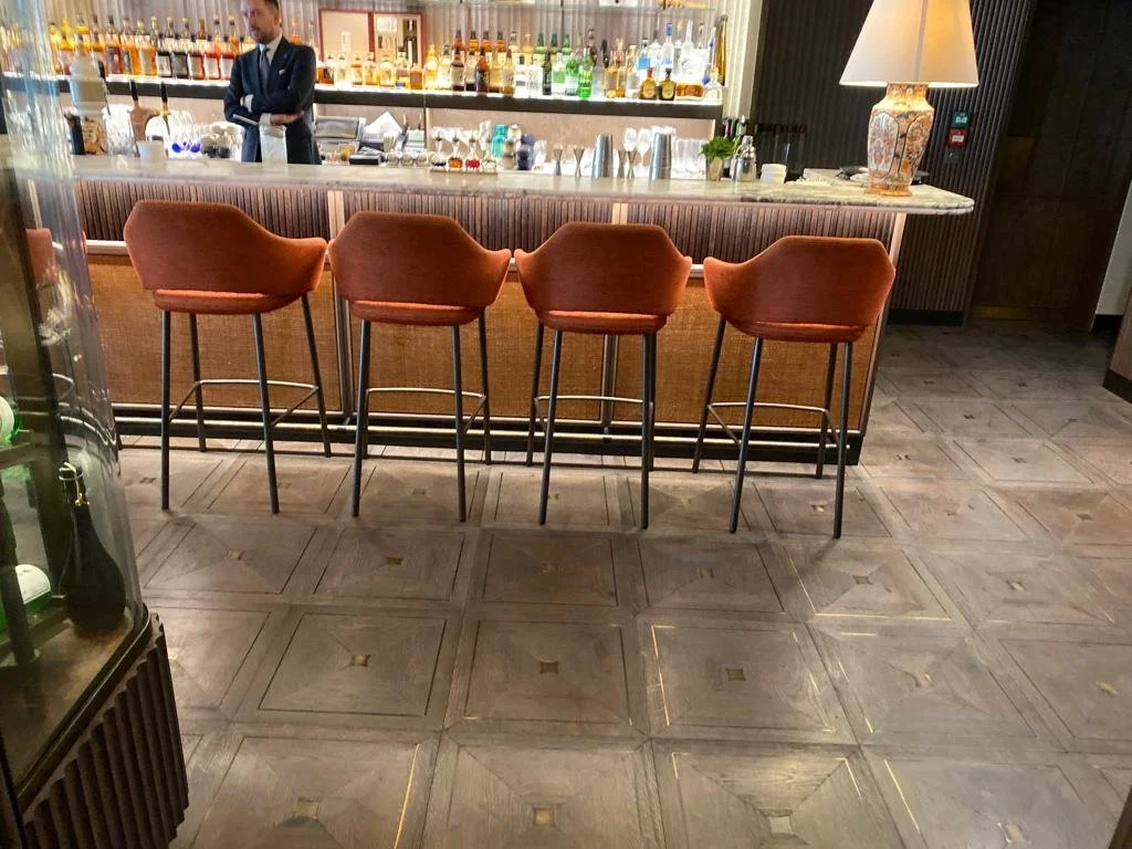 Bar con cuatro sillas acolchadas de color camel, bartender detrás del mostrador con varias botellas de alcohol y un vaso. Lámpara de mesa con base decorativa a la derecha.