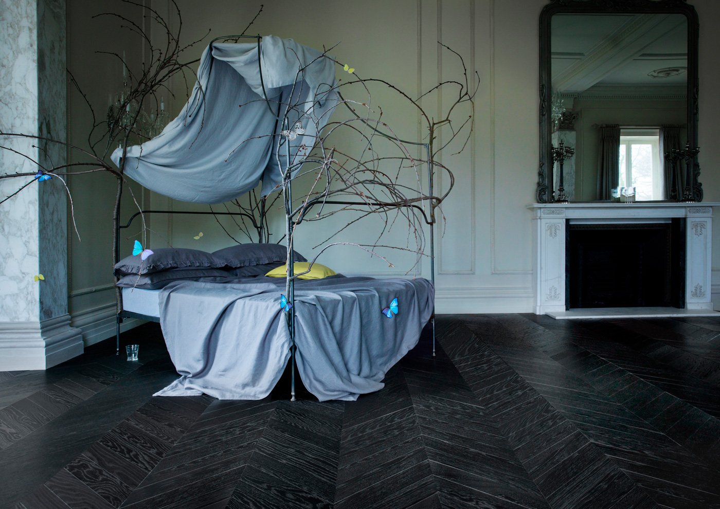 Cama decorada con ramas secas y cortinas azules en una habitación con paredes blancas y pisos oscuros de madera.