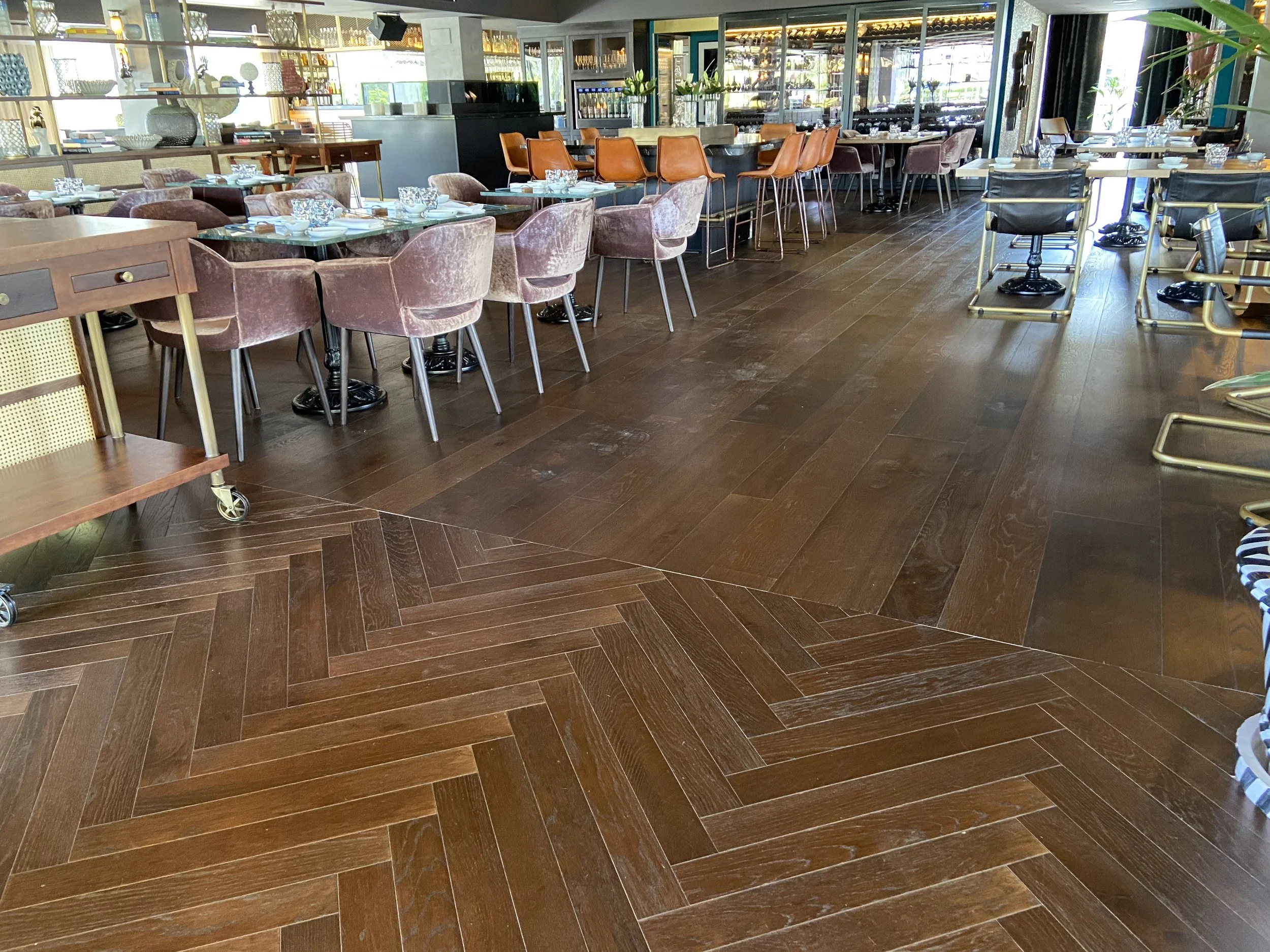 Interior de un restaurante con mesas arregladas, sillas y decoración moderna, piso de madera en diferentes patrones.