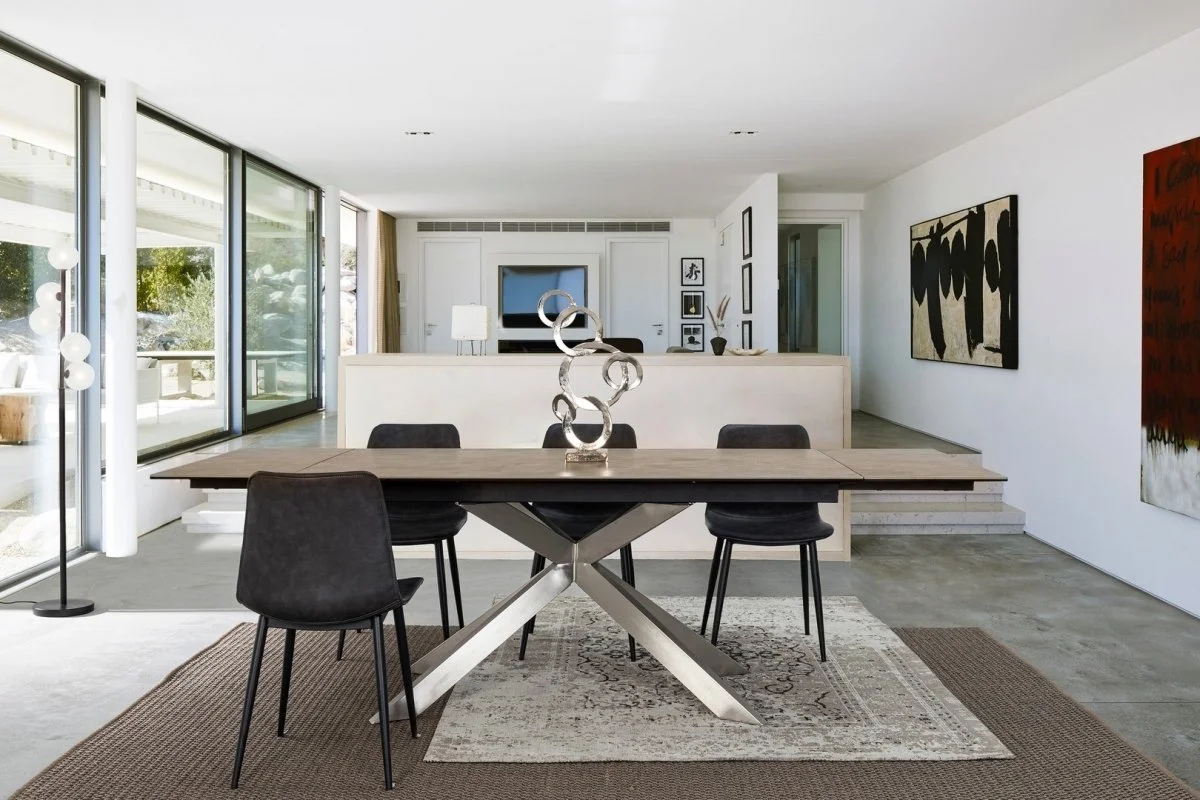 Sala de estar con decoración moderna, mesa de comedor con cuatro sillas negras, escultura de arte en el centro, paredes blancas con cuadros y gran ventana con vista al exterior