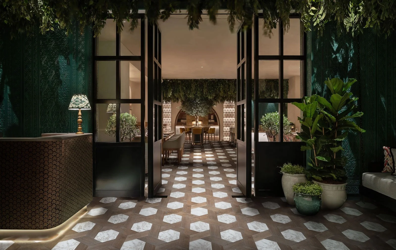 Interior de un restaurante elegante con plantas, mesas y sillas, dividido por puertas de cristal y decoraciones en tonos oscuros y verdes.
