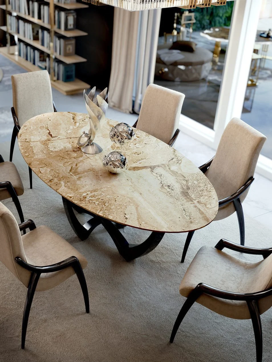 Mesa de comedor de topo de piedra con seis sillas crema con brazos oscuros, adorno decorativo en el centro y en la mesa, en una sala con vista a un patio con muebles y plantas.