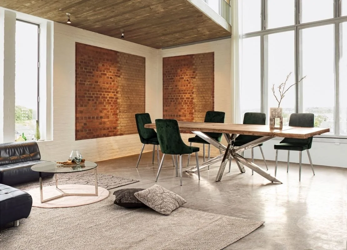 Sala de comedor con mesa de madera y seis sillas negras, paredes de ladrillo expuesto, ventanas grandes con cortinas blancas, sofá negro, mesa de centro, algunos cojines en el suelo.