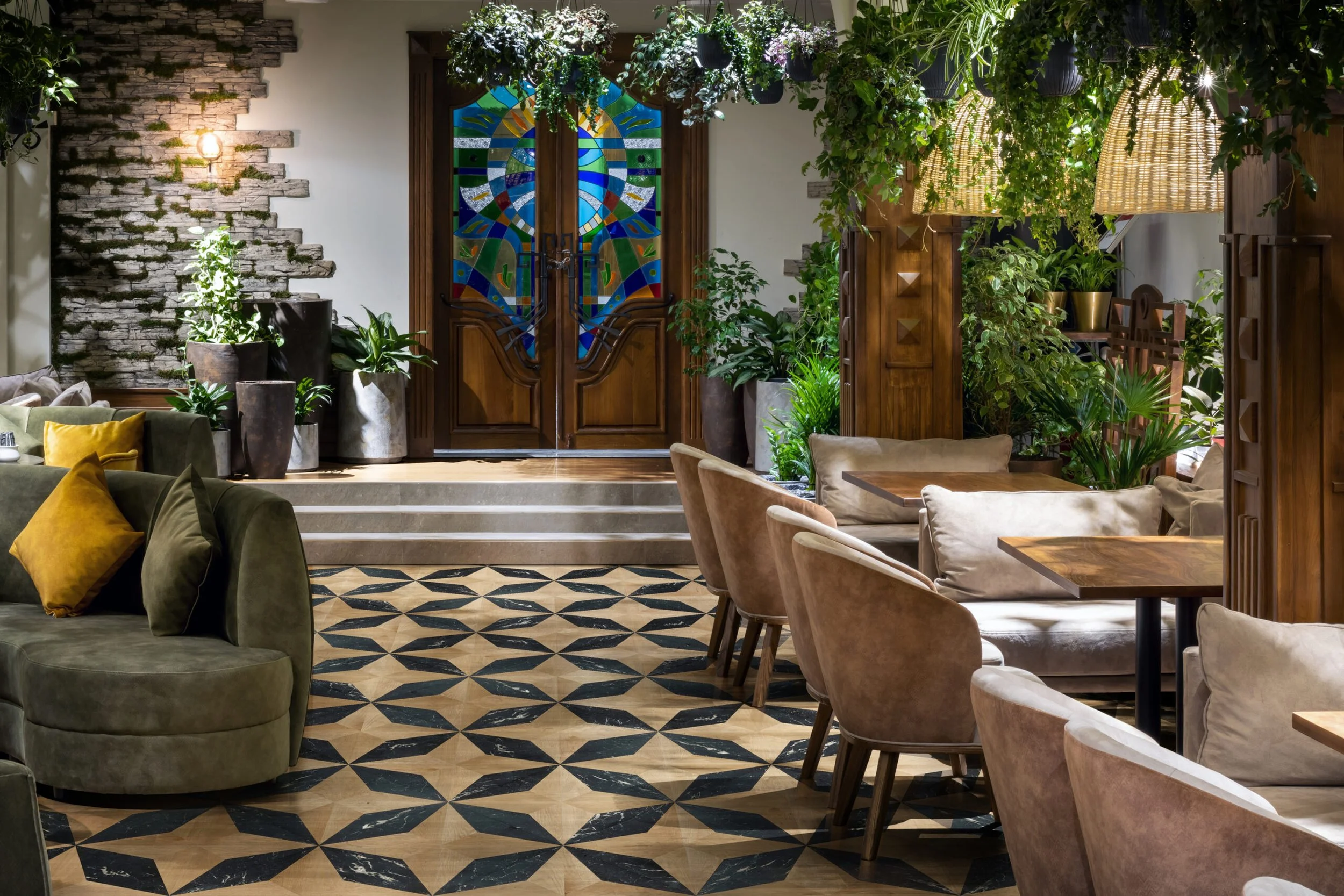 Interior elegante de un restaurante o cafetería con piso de mosaico en tonos beige y negro, muebles de madera y de colores neutros, muchas plantas verdes y lámparas colgantes de mimbre, con una puerta de madera decorada con un vitral en el fondo.