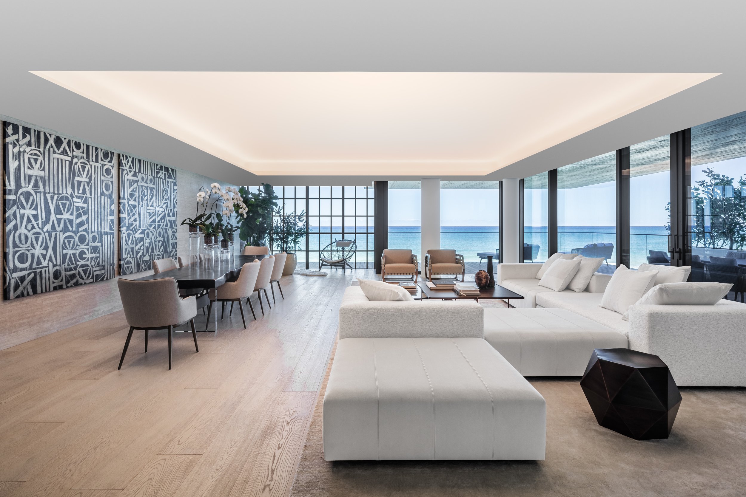 suelos de madera en salón moderno con vista al mar, sofá blanco, mesas de centro, sillones y paredes con ventanas de cristal que muestran la playa y el océano.