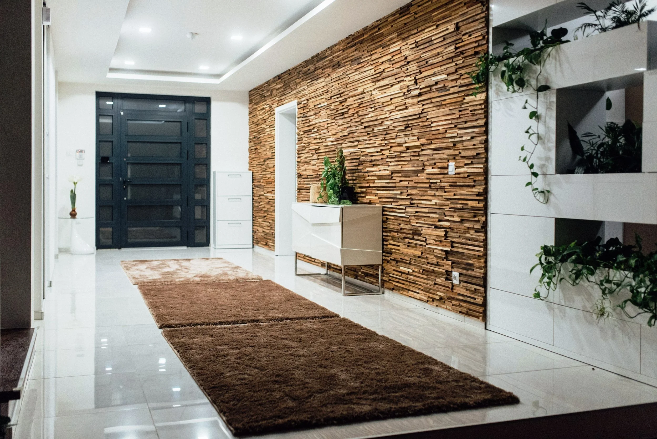 Vestíbulo con pared decorativa de madera, alfombra marrón, plantas en macetas blancas y un armario blanco junto a la pared.