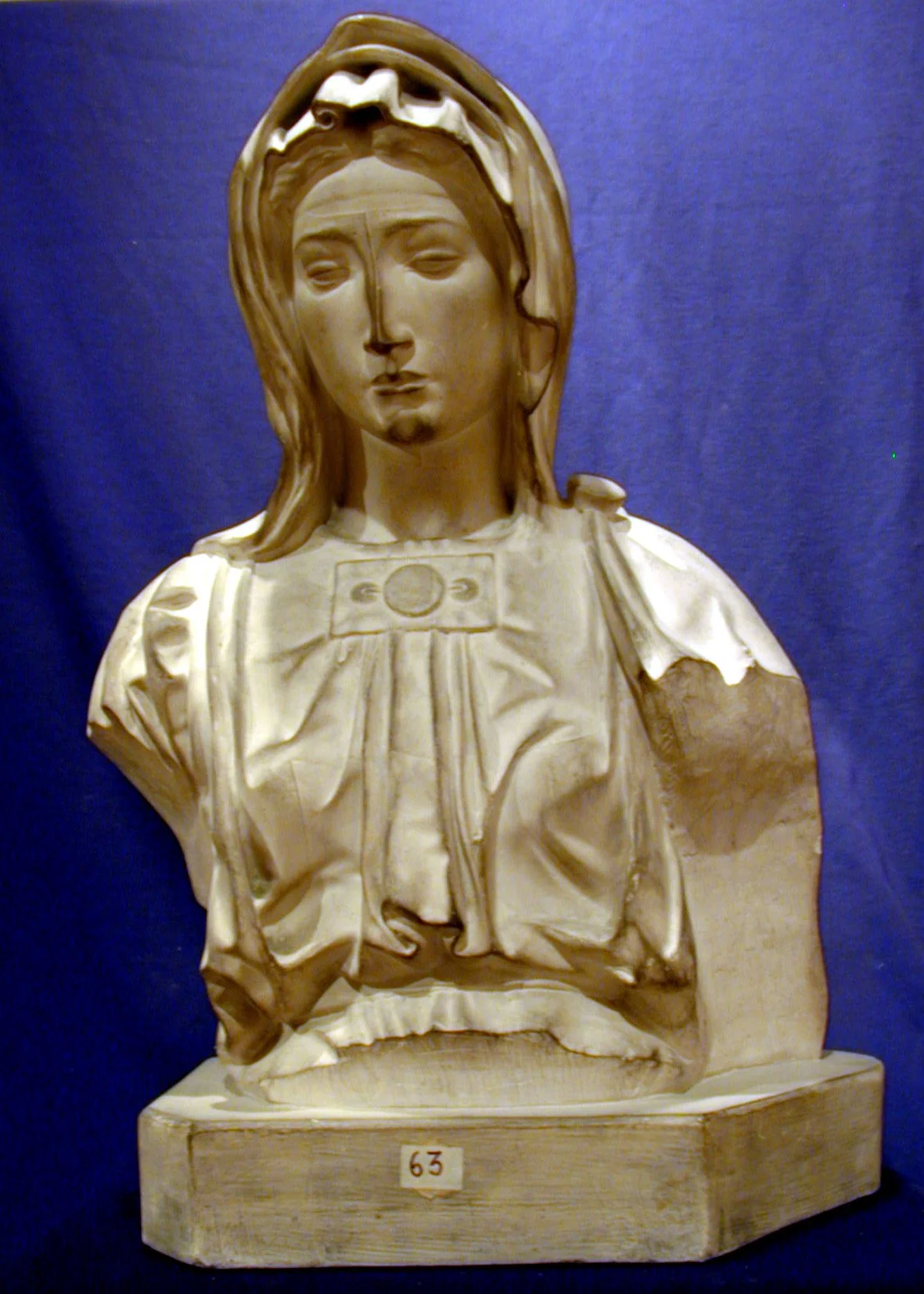 Escultura en mármol de una figura femenina con una expresión seria, con cabellera larga y cabello con detalles. La escultura muestra detalles faciales y vestimenta antigua. Está colocada sobre una base de madera marcada con el número 63.